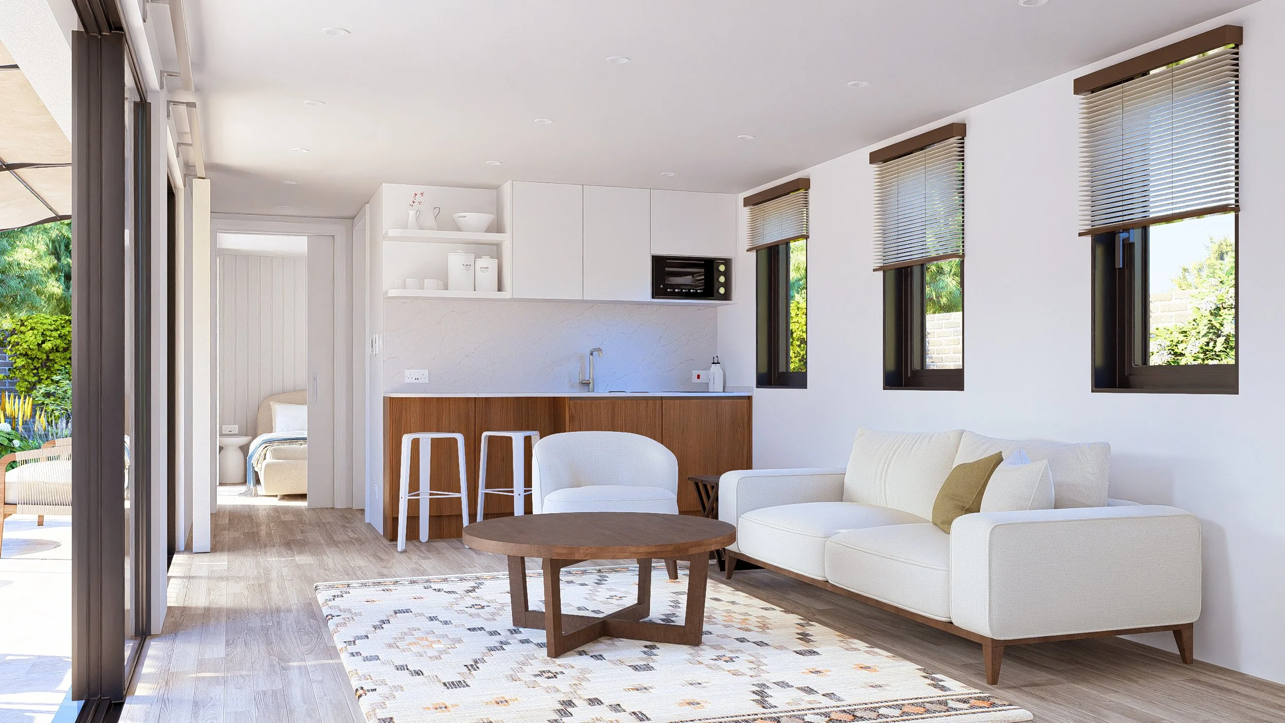 Bauhu hurricane resistant modular hospitality p living room with white sofa, wooden coffee table, and three windows with blinds. Open kitchen with white cabinets and wooden accents, bar stools, and door leading to a bedroom with a bed and side table.