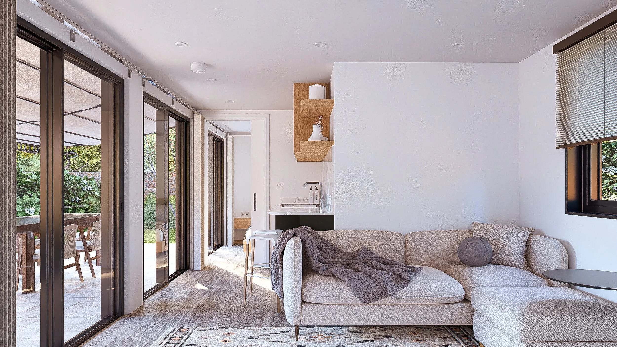 A modern living room with sliding glass doors opening to an outdoor patio with a dining table and greenery. The room features a light-colored sectional sofa with a blanket and a round pillow, a small side table, and a window with blinds. The kitchen 