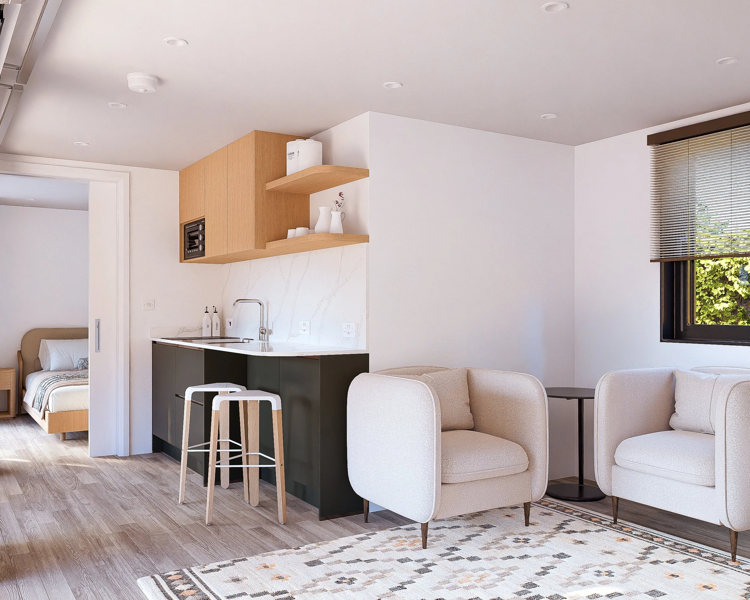 Open-concept living space with a small kitchenette, two white armchairs, a black side table, a patterned area rug, and a bedroom visible through a doorway. The room has light wood flooring, white walls, and a window with blinds.