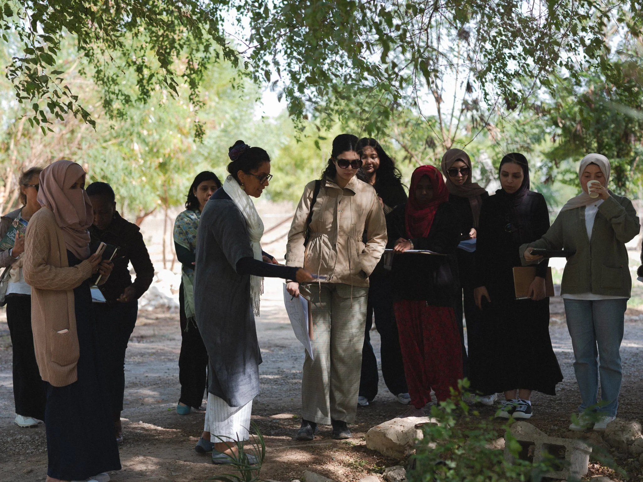Group of women standing outdoors under trees, some holding notebooks and looking at a woman showing something on a piece of paper. One woman is drinking from a cup.