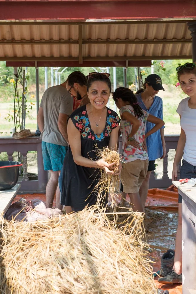A group of people in an outdoor barn-like shelter, with a woman in the center smiling and holding a bunch of straw, surrounded by others preparing for a traditional pig slaughter. The scene is rural with trees and open land in the background.