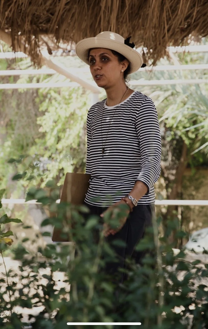 A woman wearing a wide-brimmed hat, striped shirt, and jewelry standing outdoors amidst lush greenery and foliage, possibly in a garden or park.