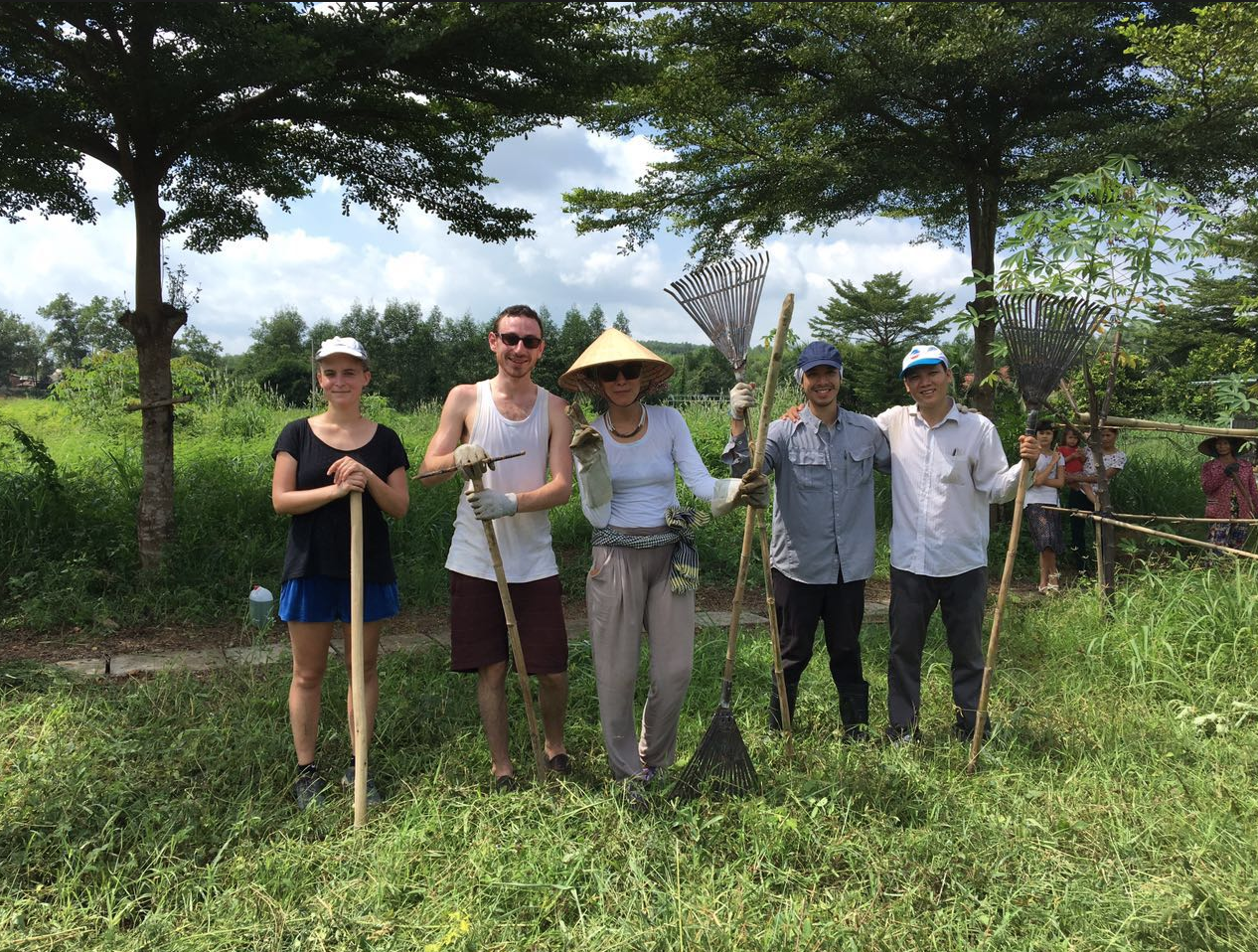 Group of five people standing outdoors with gardening tools, smiling, surrounded by trees and grass, with a few people in the background.