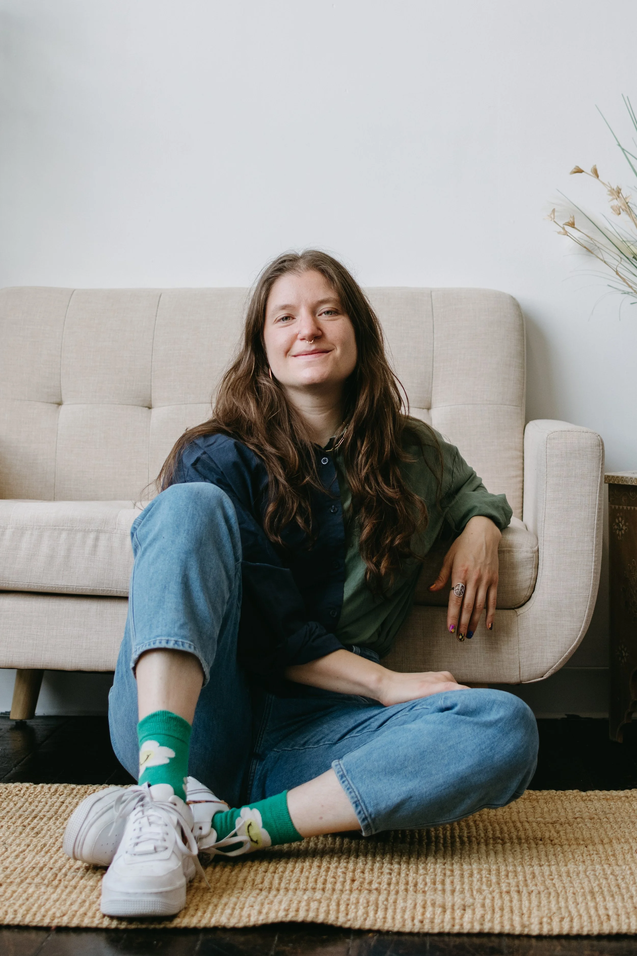 Sarah sits on the ground in front of a tan couch. She is wearing blue jeans and a blue and green button up shirt. Sarah is smiling and has long, curly brown hair.