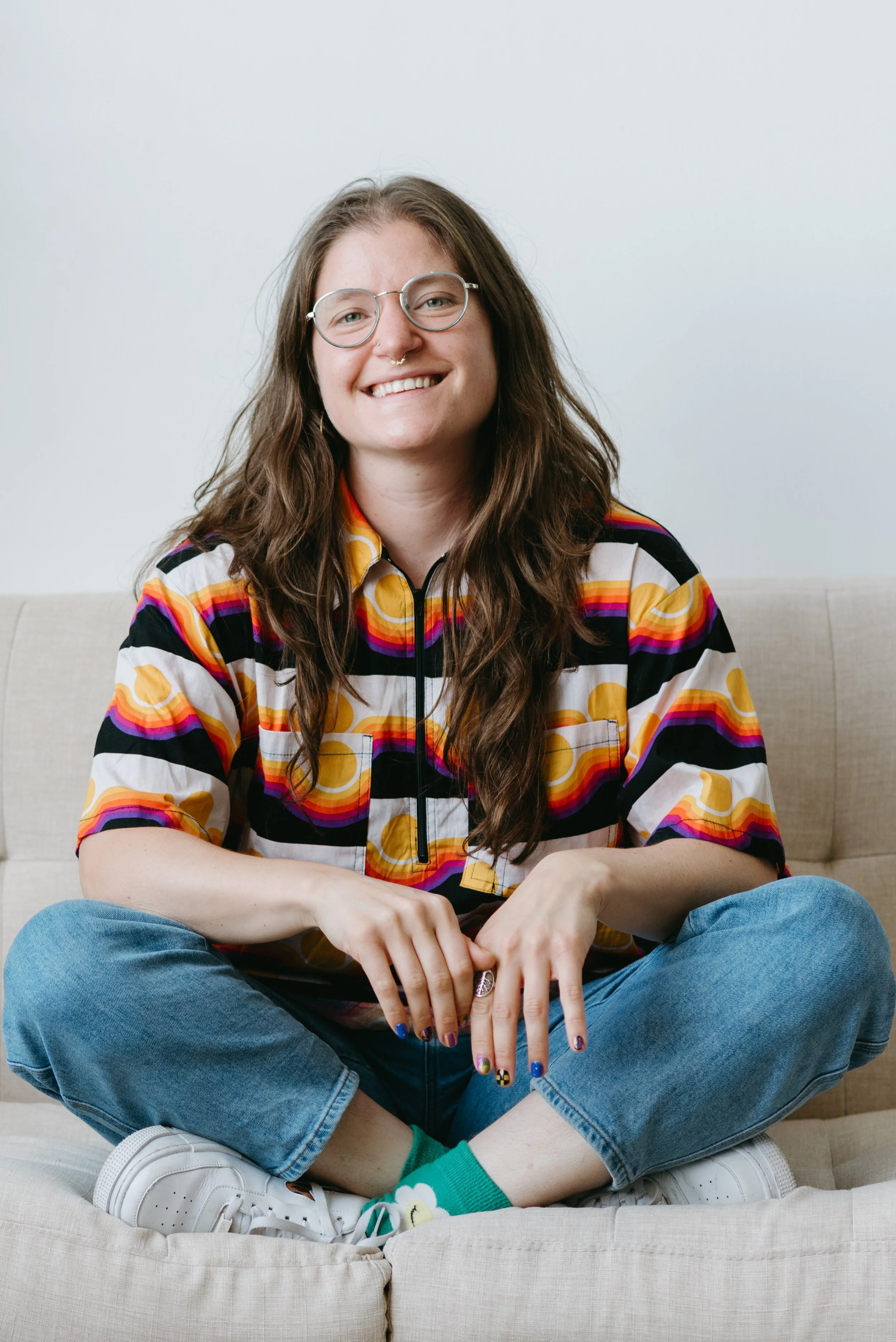 Sarah Marcus, LCSW, a white person with brown, wavy hair, sits crosslegged on a tan couch smiling. She is wearing wire-rimmed glasses, a yellow, purple, and red patterned shirt, and blue jeans.