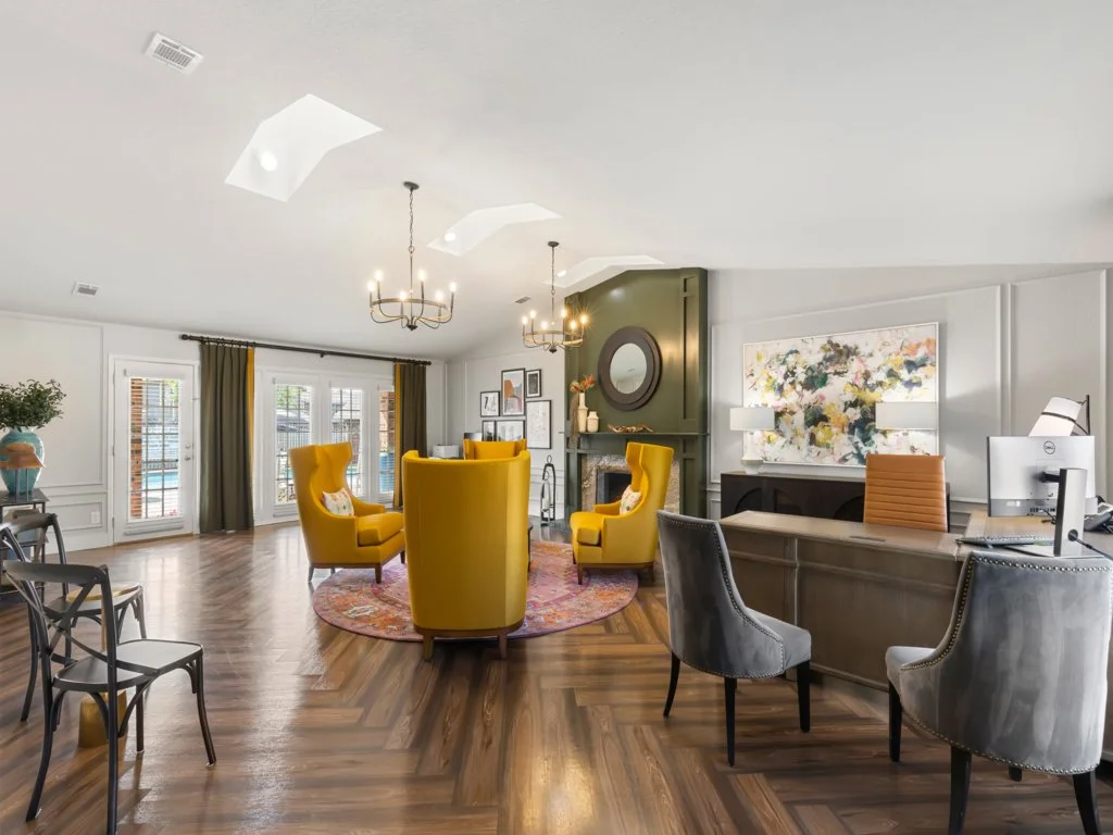 Townhome lobby with four yellow armchairs arranged around a circular area rug, a fireplace with a round mirror above, gray and orange chairs near a desk with a computer, and large windows with dark curtains, all on hardwood floors.