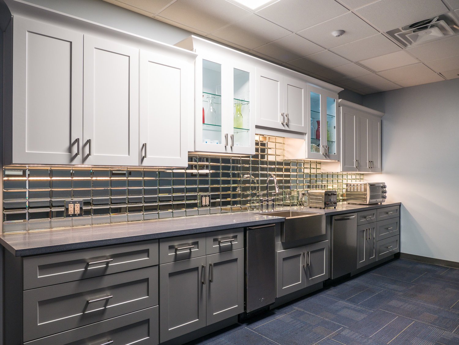 Kitchen with gray lower cabinets, white upper cabinets, a dark countertop, a black and gold tiled backsplash, a sink with a high faucet, and small appliances on the counter.