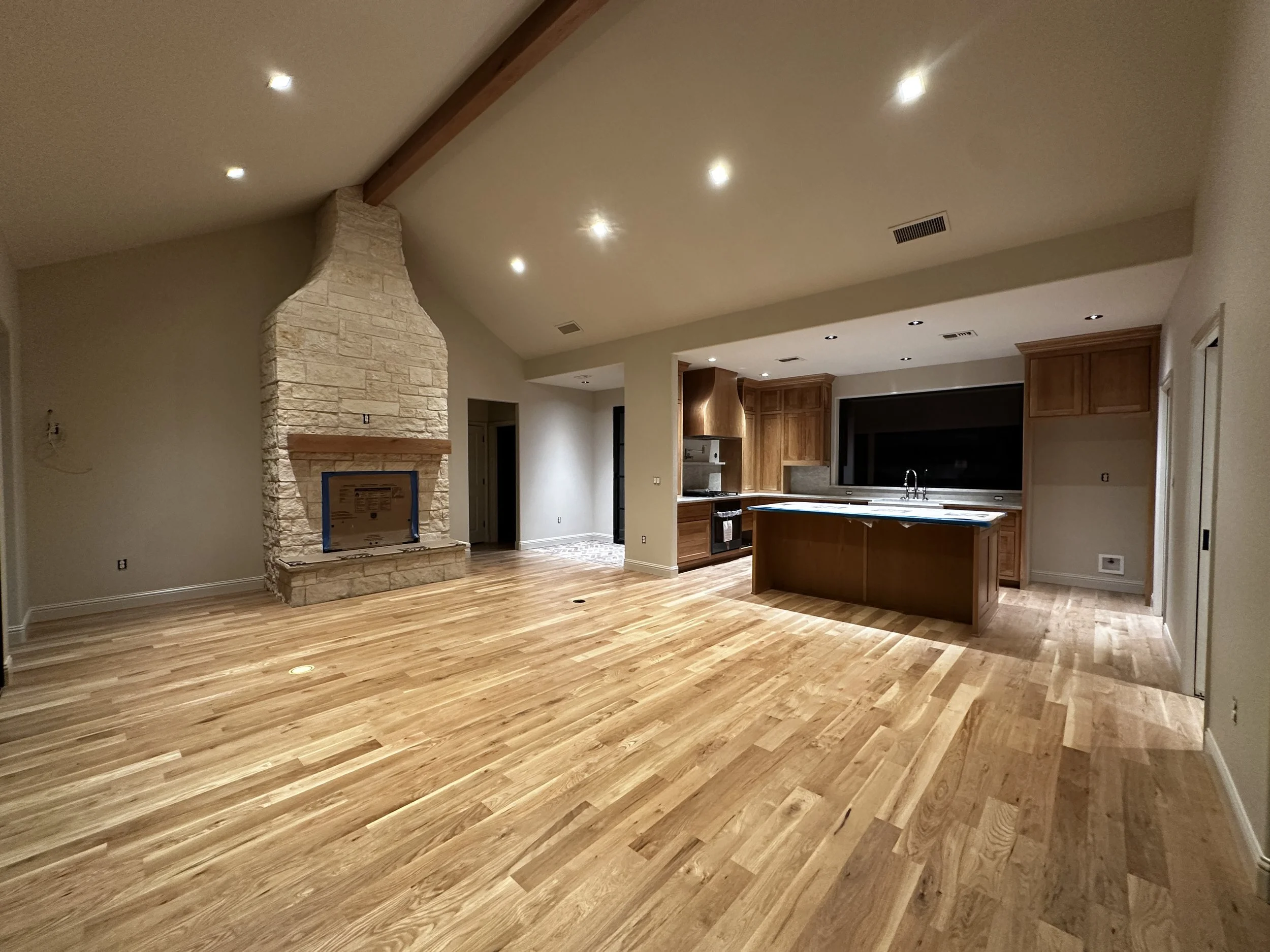 Spacious open living area with hardwood floors, a stone fireplace, and a kitchen with wooden cabinets and a central island.