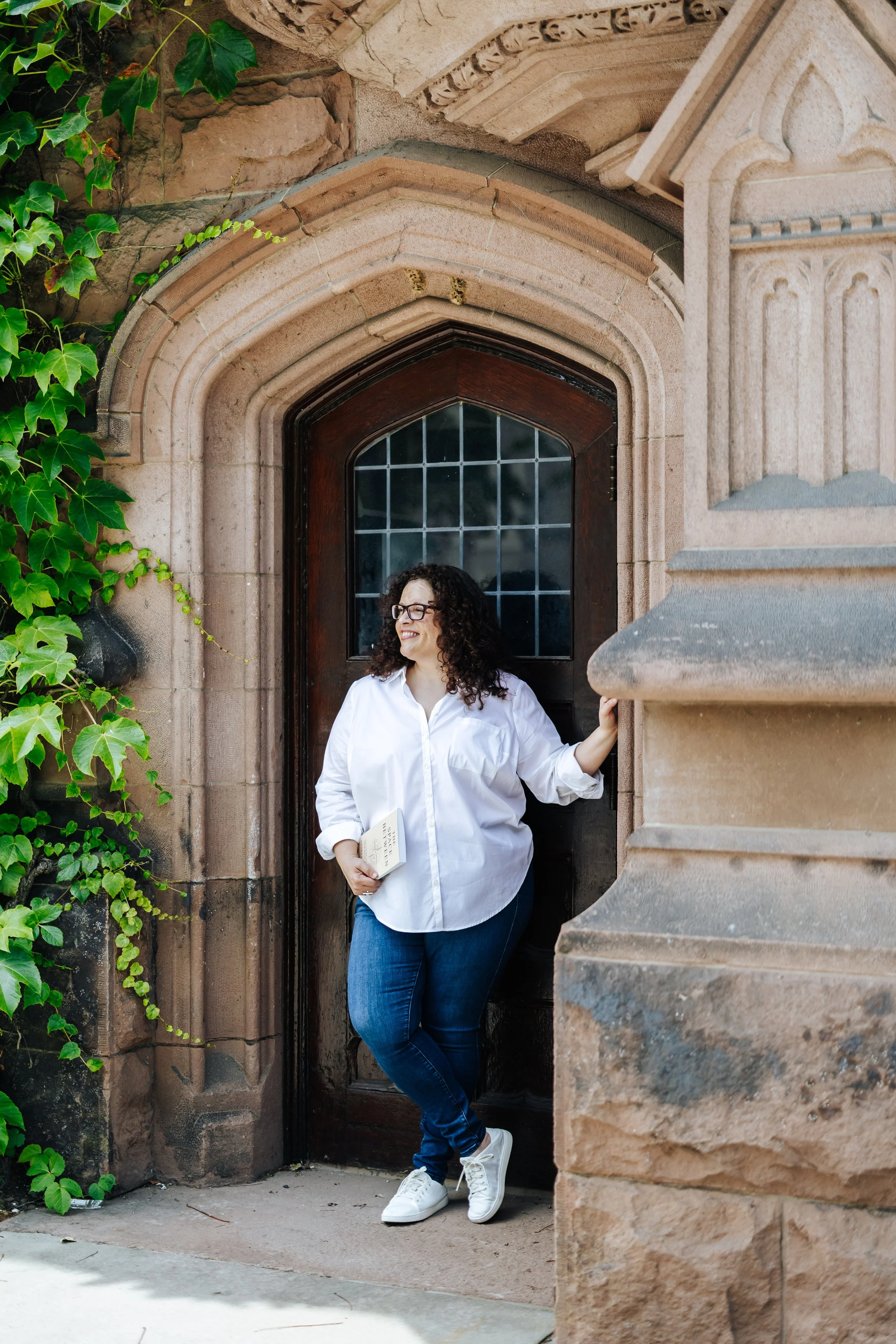A woman with curly hair and glasses, wearing a white shirt and jeans, standing in a doorway of a historic building with ornate stonework, holding a book in her left hand and smiling.