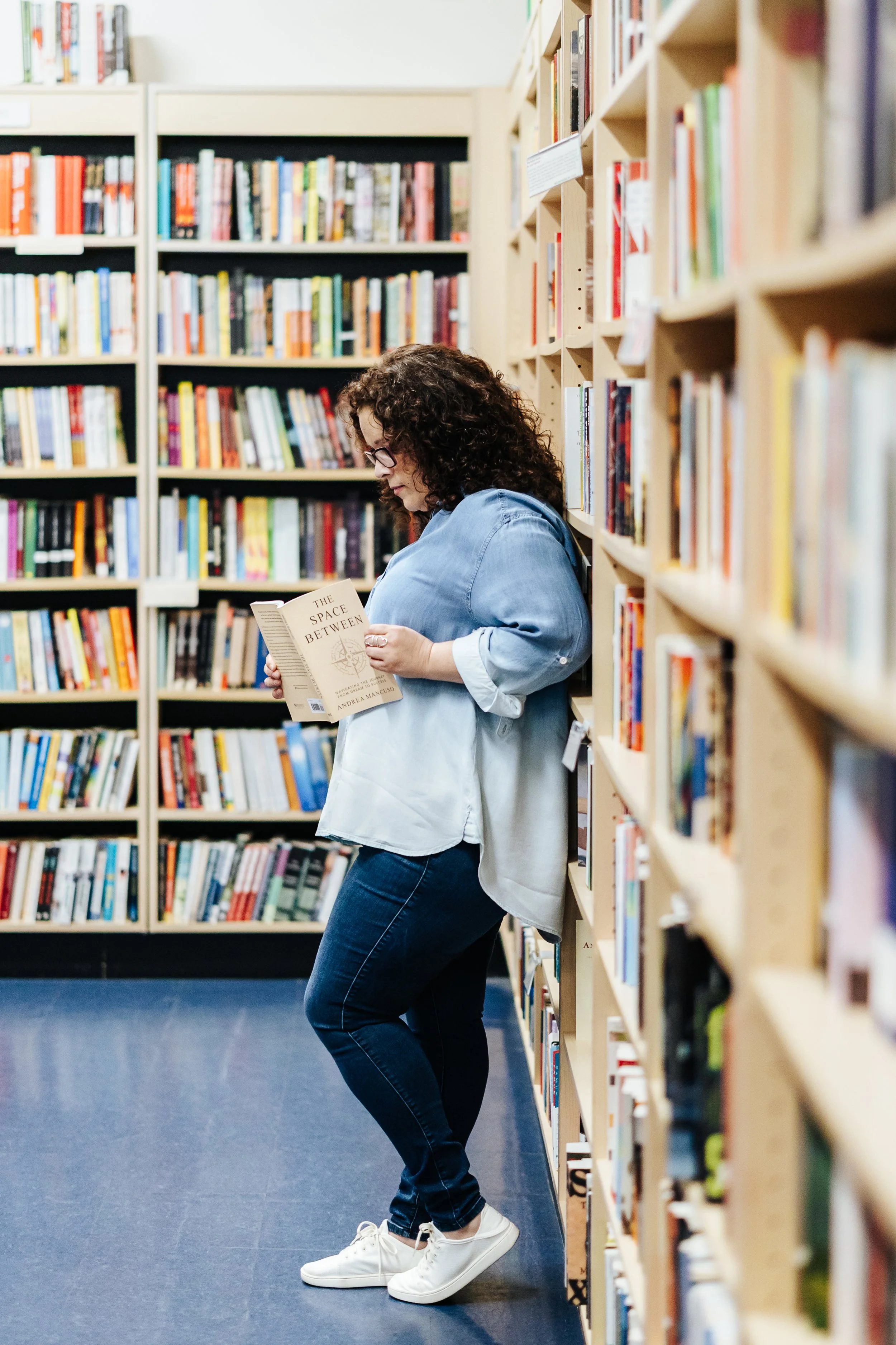 A woman with curly hair and glasses leaning against a bookshelf, reading a book titled 'The Space Between' in a bookstore or library.