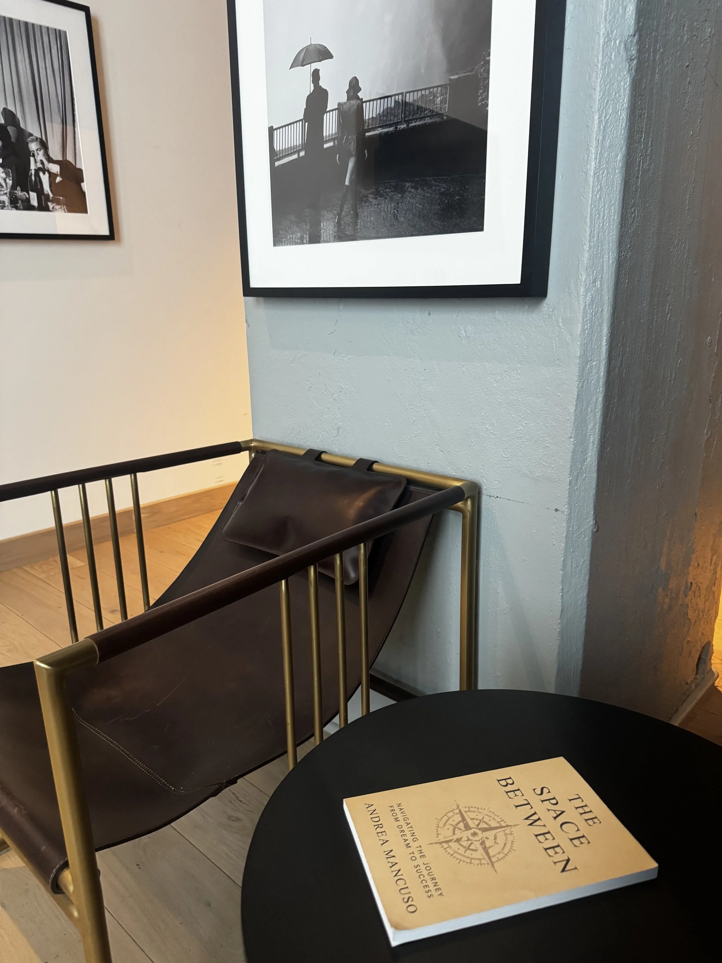 A black book titled 'The Space Between' resting on a black round table in an art gallery, with a black leather chair with gold-colored metal armrests and a framed black and white photograph on the wall behind it.