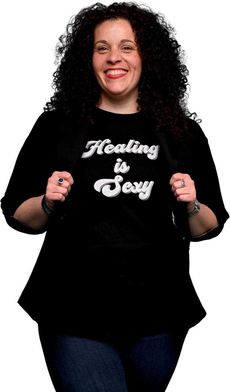 A woman with curly hair smiling and holding open her jacket to reveal a black t-shirt that says 'Healing is Sexy' in white cursive text.