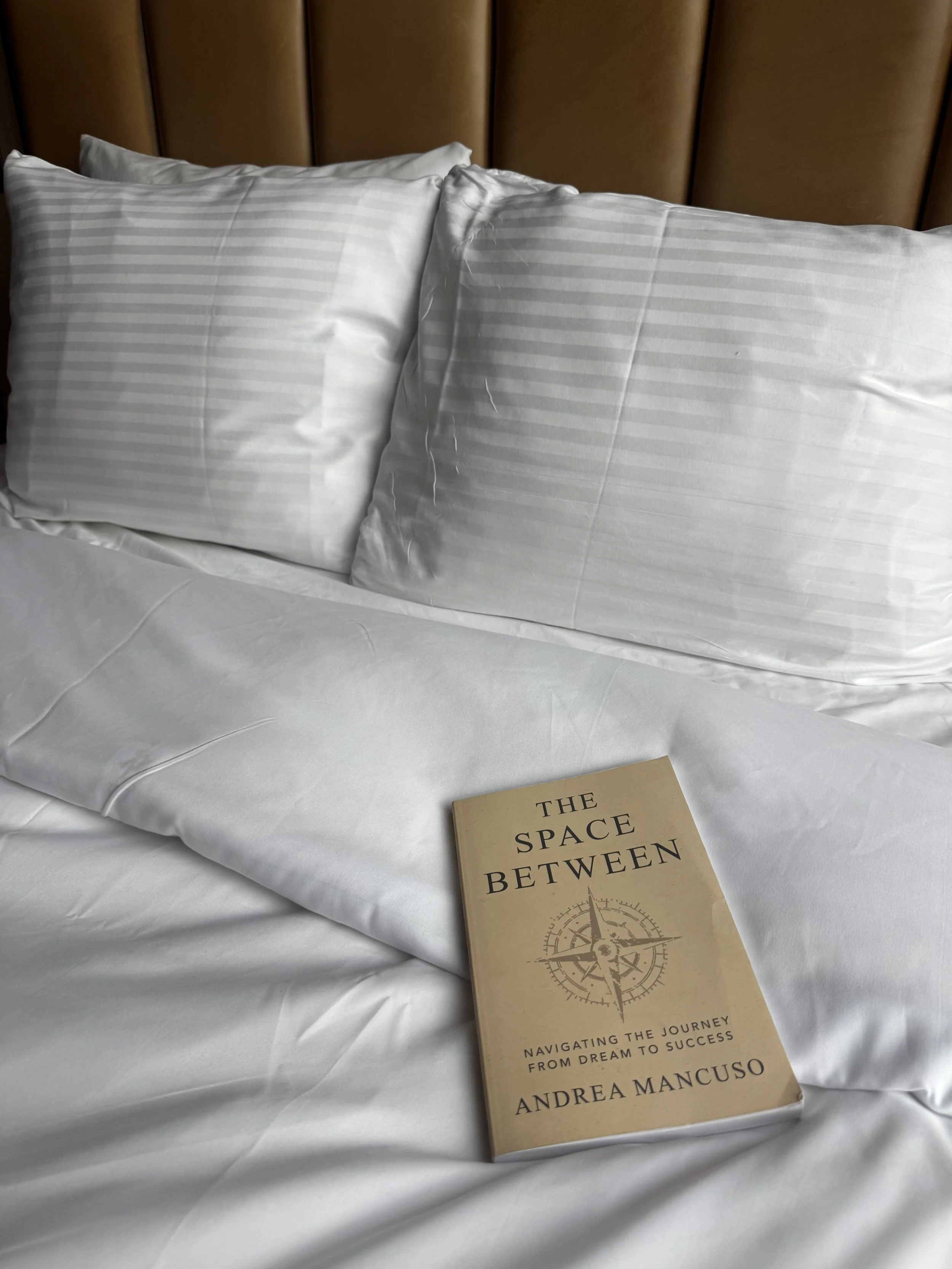 A bed with white striped pillows and a beige book titled "The Space Between" resting on the white bedspread. The headboard is made of brown leather or leather-like material.