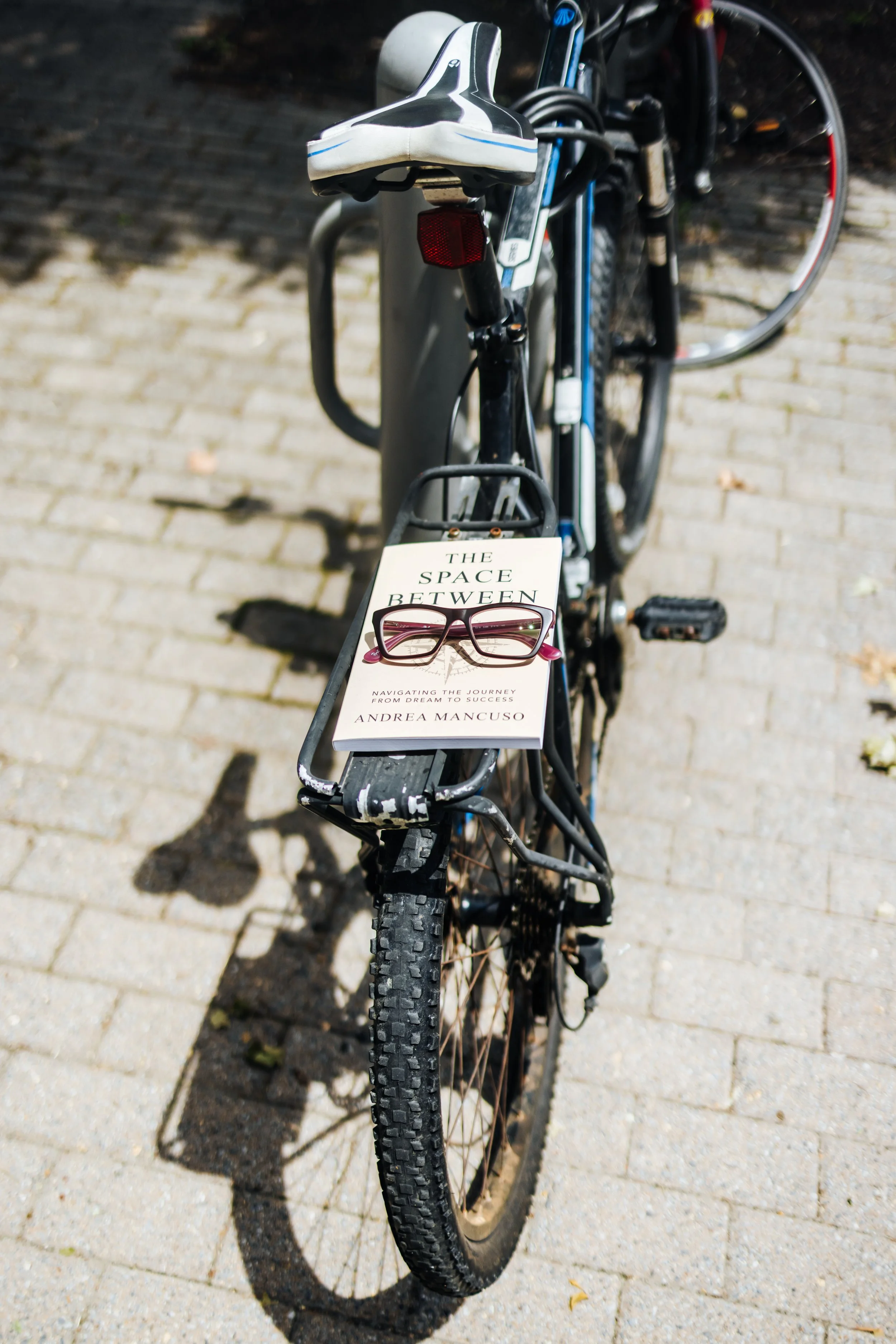 A black mountain bike parked on a brick pathway, with a white book titled 'The Space Between' and a pair of glasses resting on the handlebars.