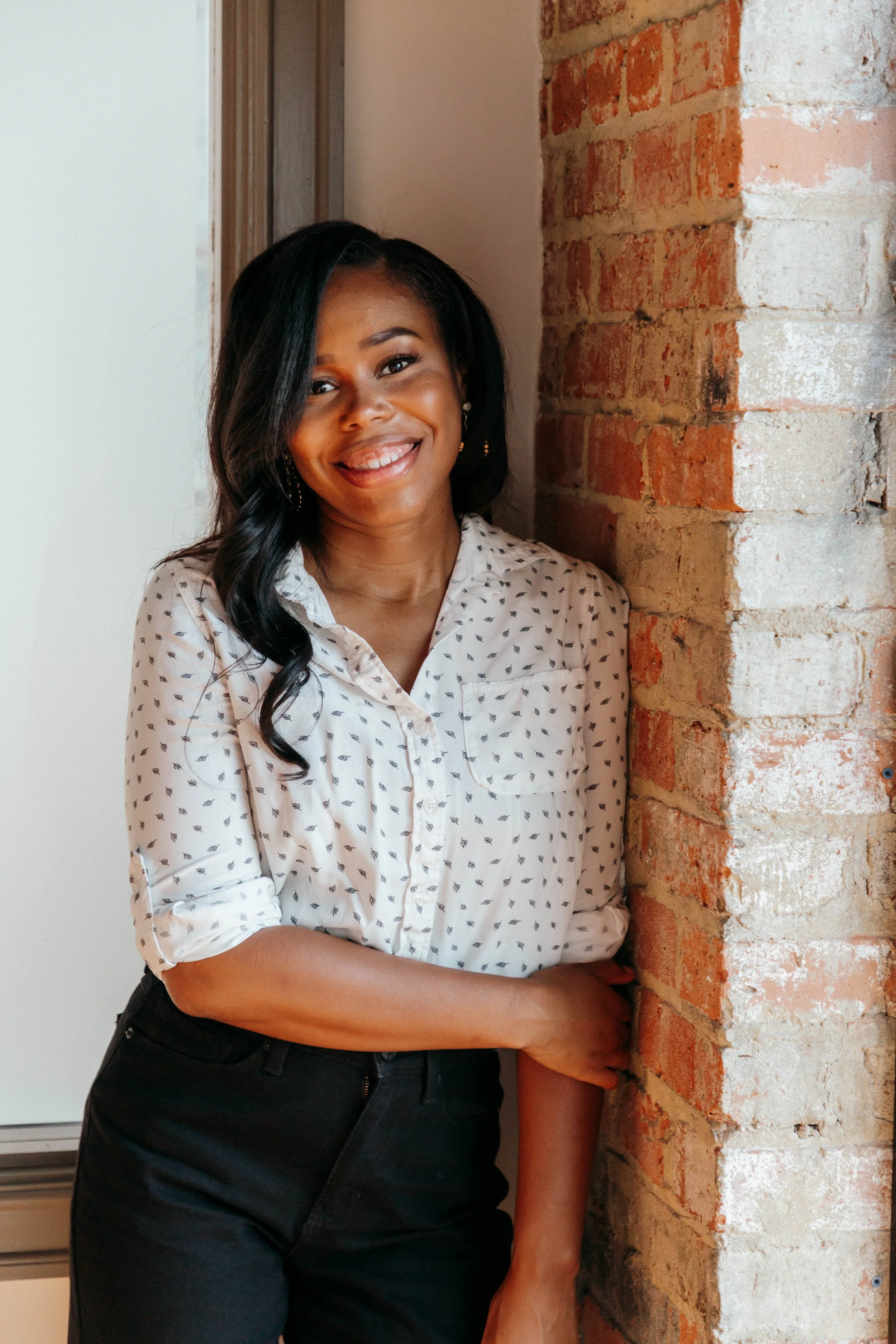 A woman with dark hair, wearing a white blouse with small black patterns, black pants, is smiling and standing next to a brick wall.