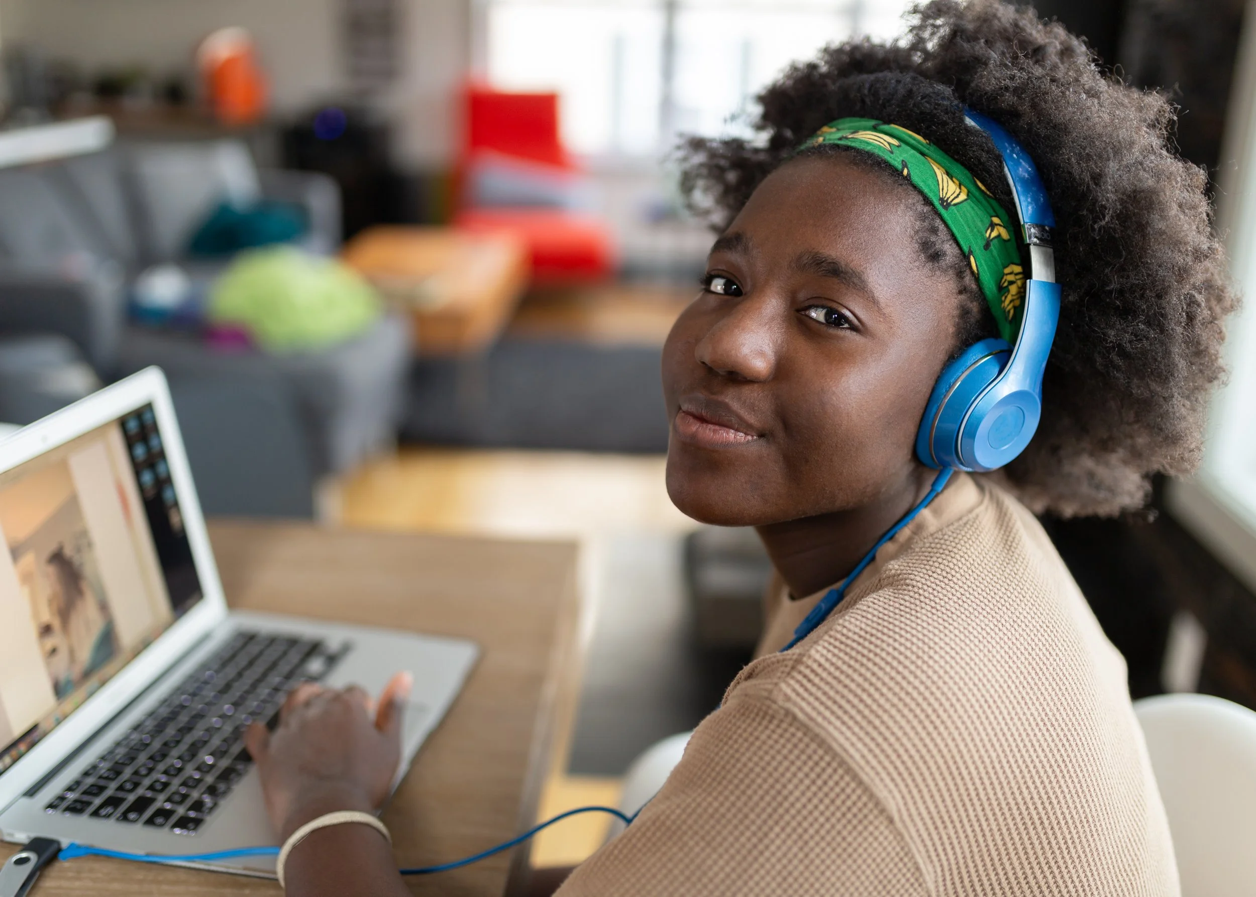 A young girl with curly hair wearing blue headphones and a green headband with banana print, sitting at a desk using a laptop.