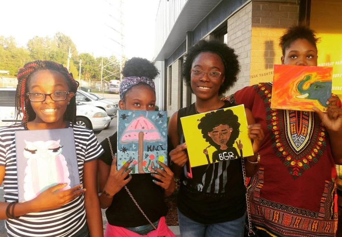 Four girls standing outside, each holding a painted canvas with colorful artwork.