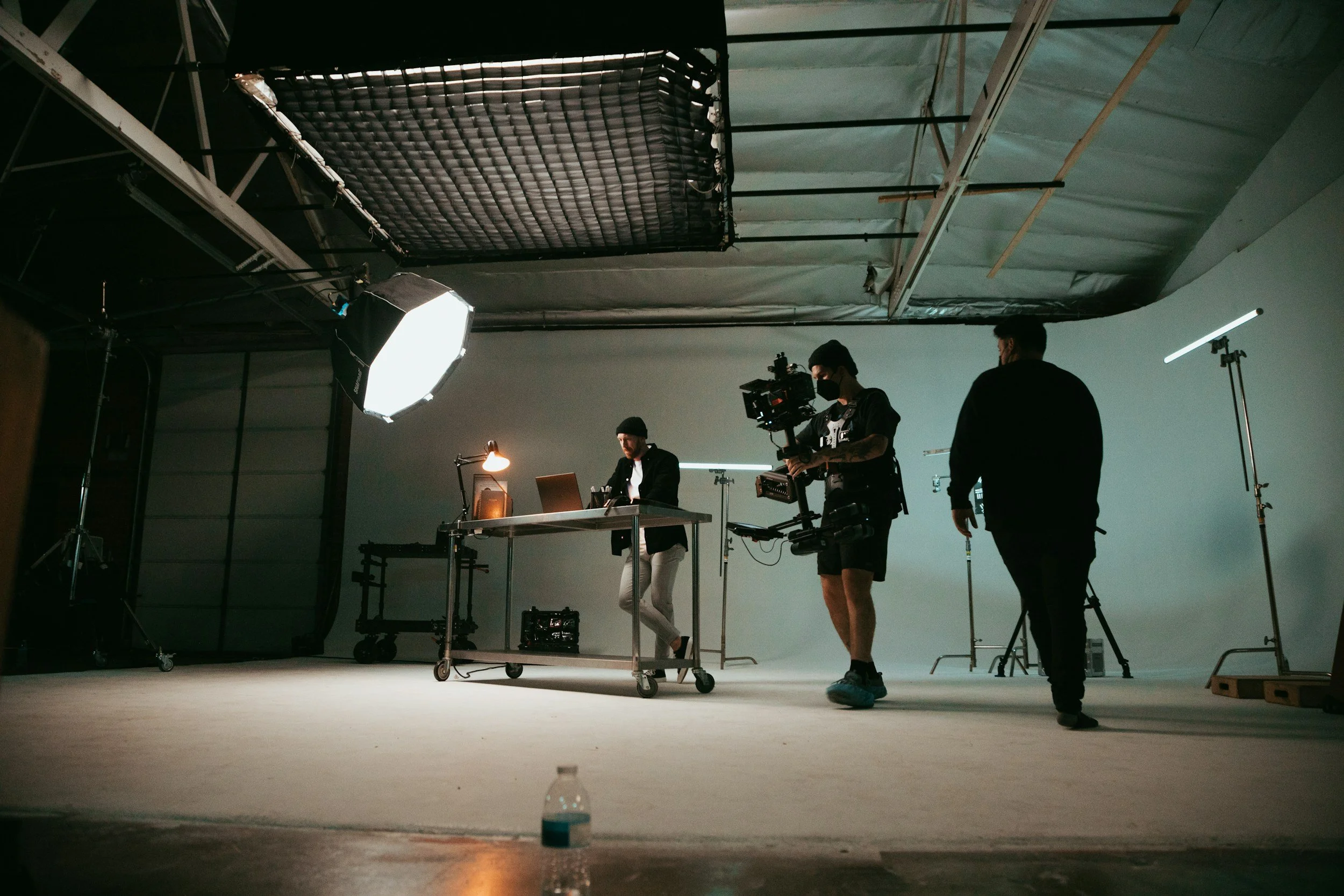 A behind-the-scenes photo of a film or photo shoot studio with crew members setting up equipment, including lights and cameras, in front of a large blank backdrop.