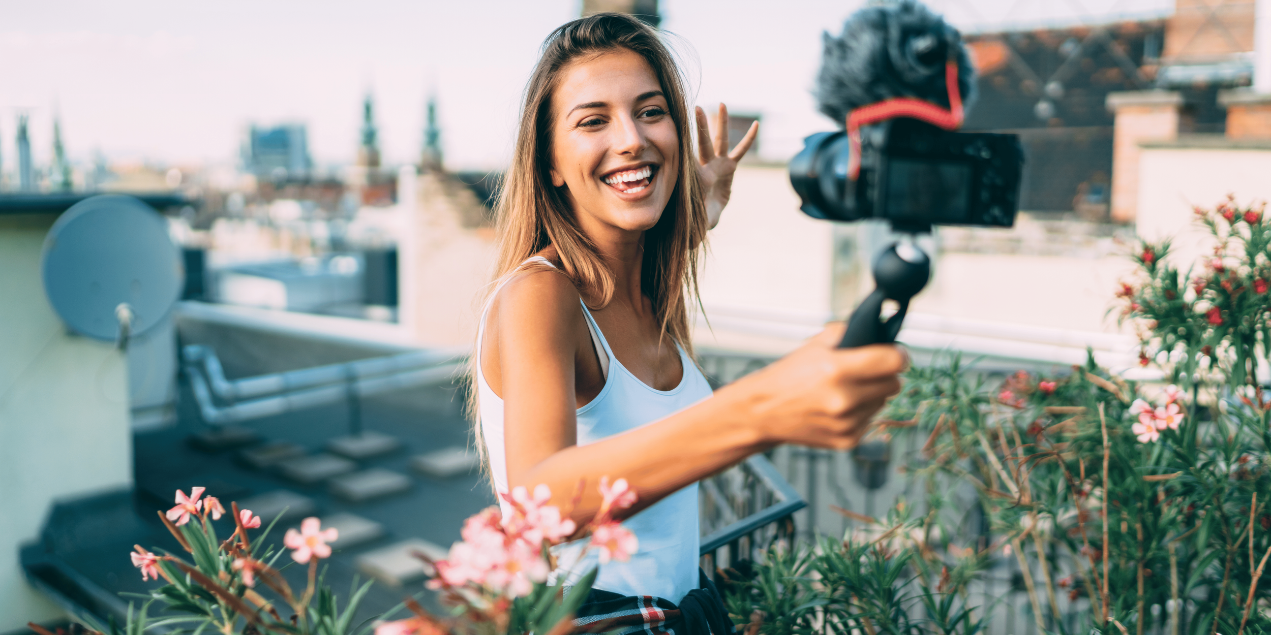 Smiling female influencer filming vlog outdoors: A young woman in a white tank top films herself with a handheld camera on a rooftop surrounded by flowers, capturing lively travel and lifestyle content.