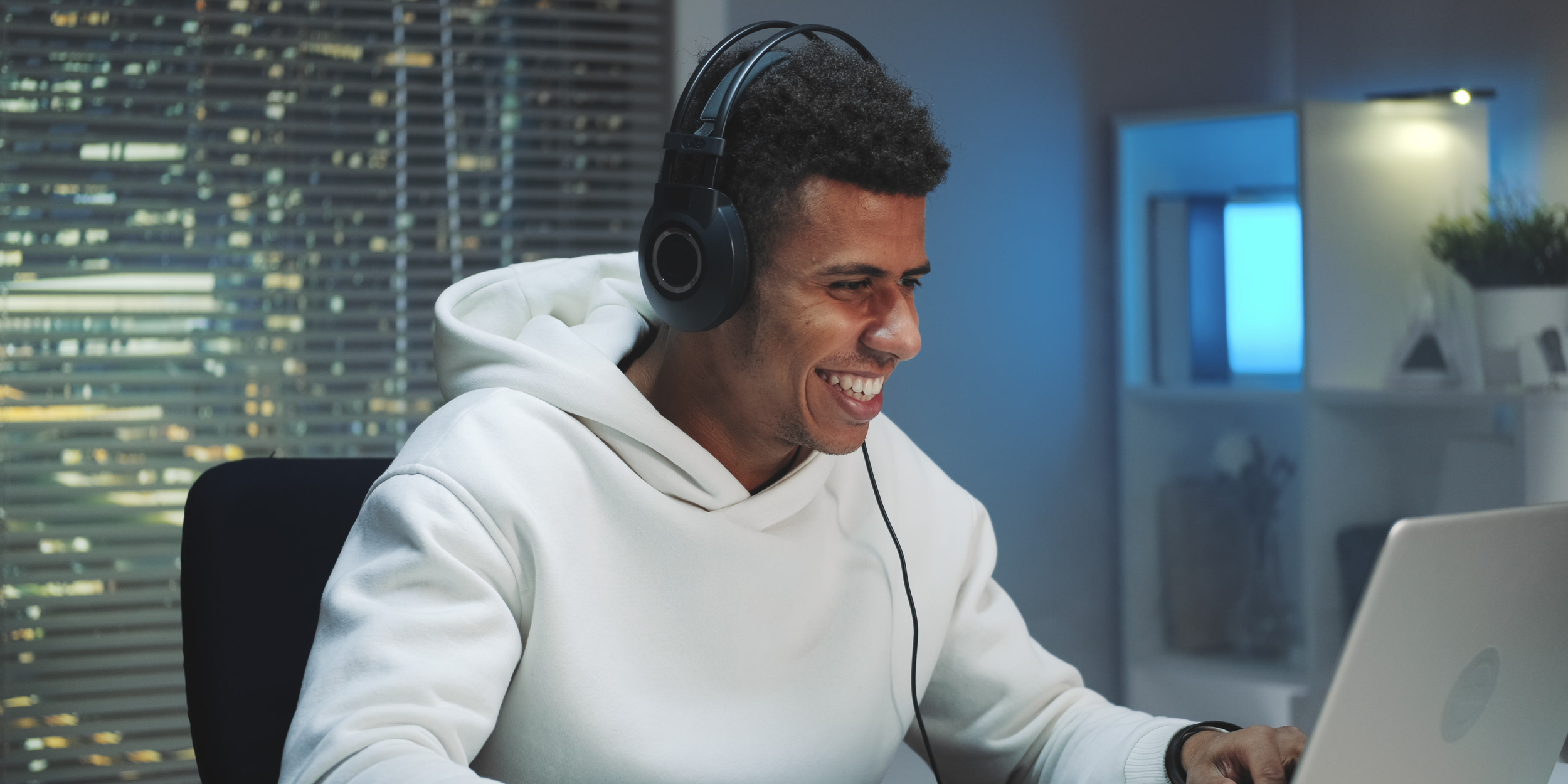 Male gamer streaming content at night: A smiling young man wearing headphones and a white hoodie plays on his laptop, creating engaging gaming or tech-related content for his audience.