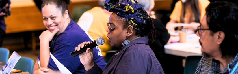 A person with medium, complexion a blue and gold head wrap glasses and a purple jacket is speaking into a microphone in the background several people appear to be listening