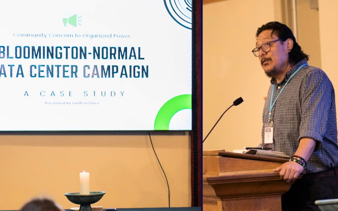 A man with a light caramel complexion and dark hair is standing at a lectern there is a video screen in the image that says Bloomington Normal Data Center Campaign a case study in the lower left there is a candle