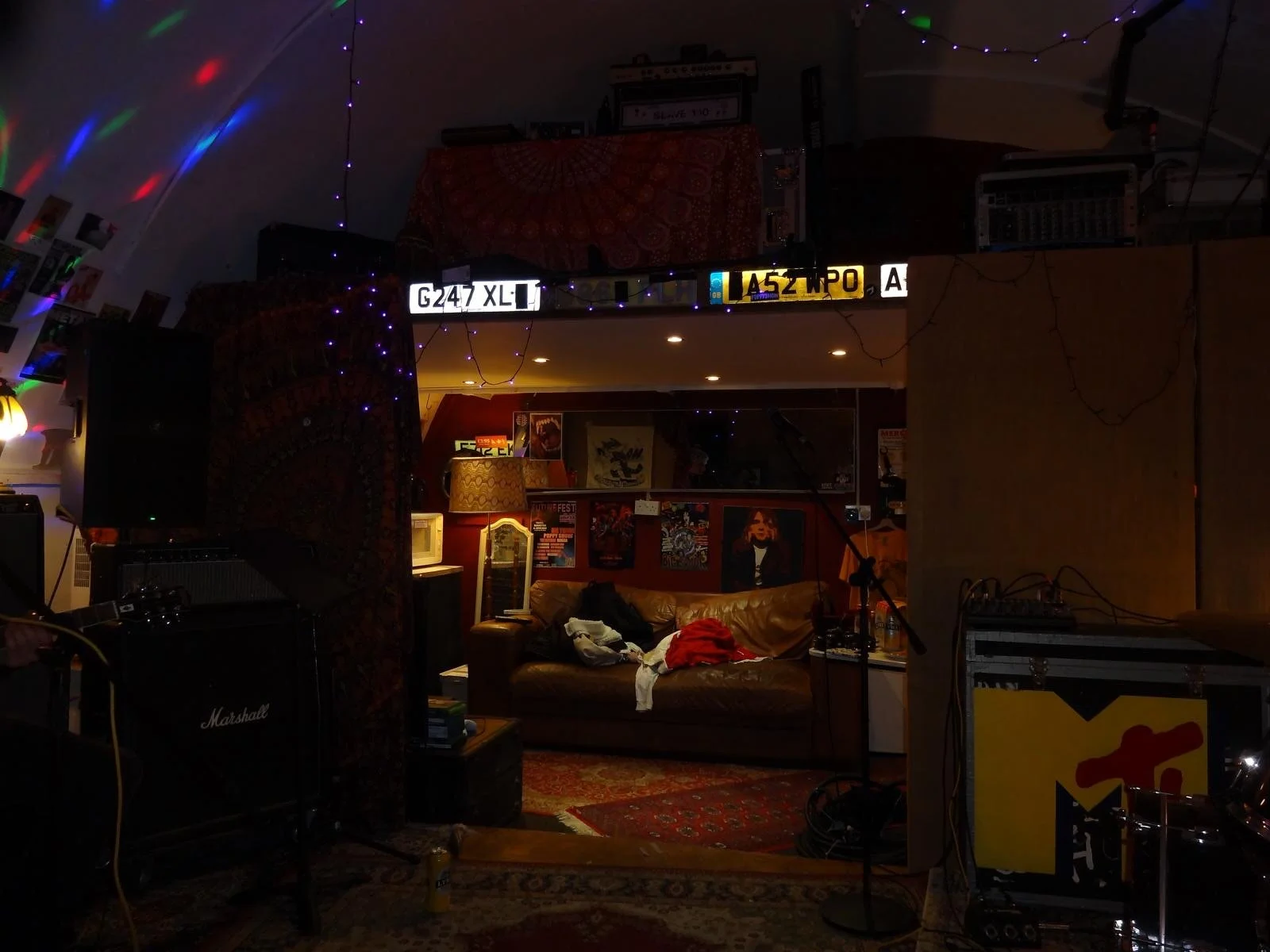 Dimly lit music or rehearsal room with a leather sofa, posters on the wall, and musical equipment including a Marshall amplifier and microphone stand, illuminated by soft warm lighting and colorful decorative string lights.