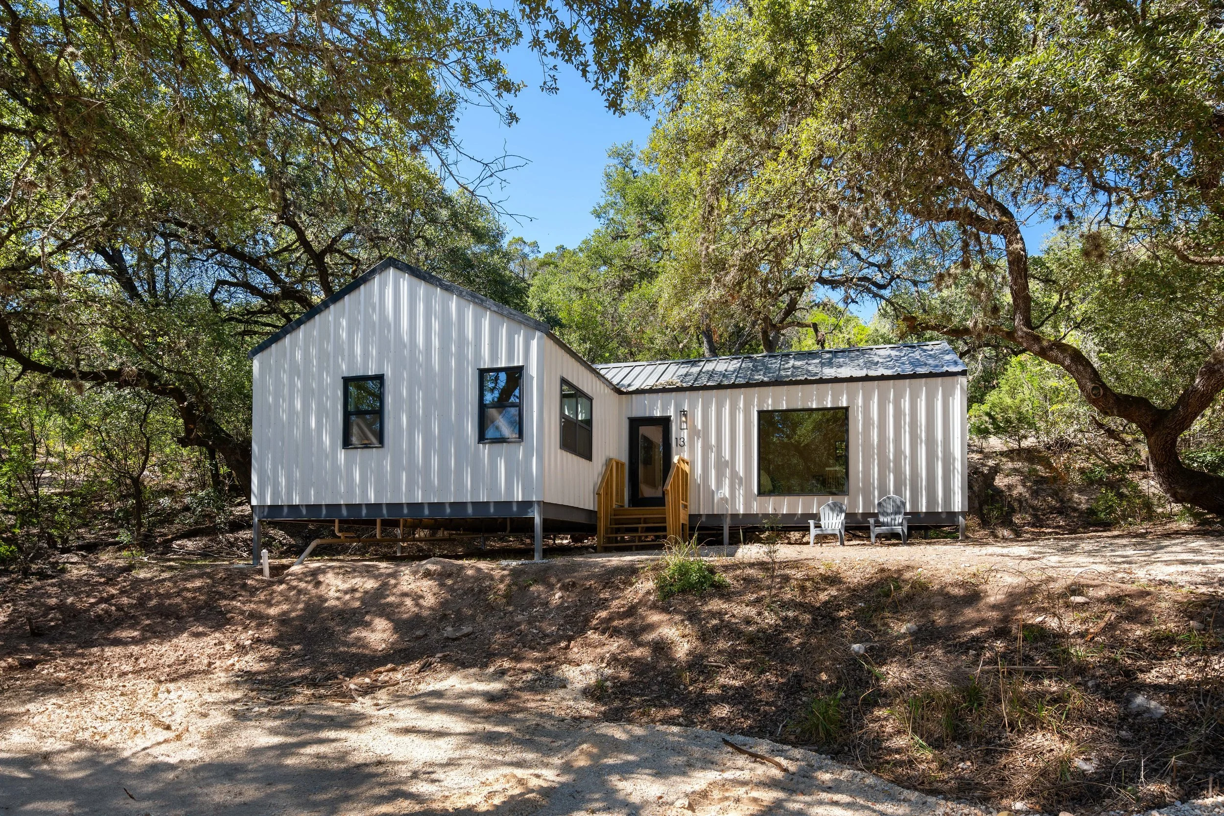 A modern white metal house on stilts surrounded by trees, with a small wooden staircase leading to the front door and two white outdoor chairs on the ground outside.