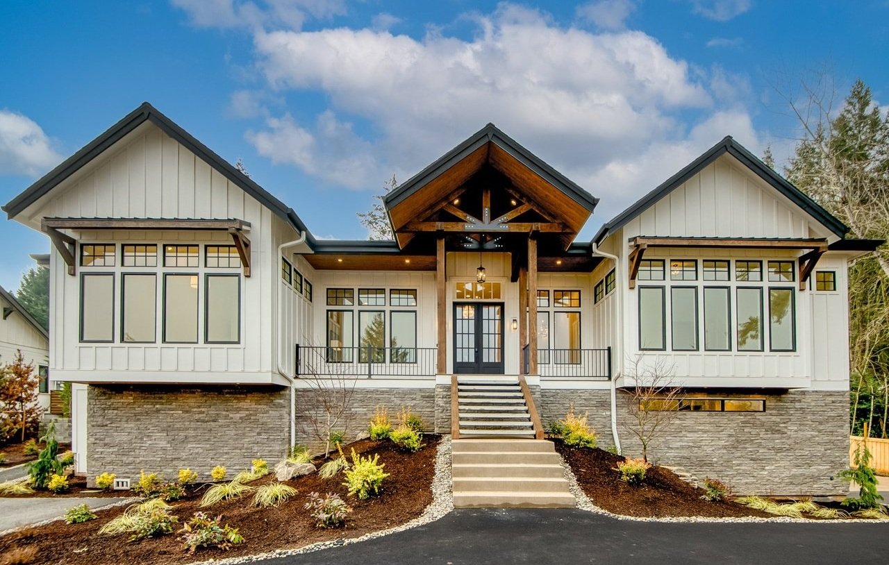 Modern two-story house with white siding, large windows, stone foundation, front porch with steps, surrounded by landscaped garden and trees, under a partly cloudy sky.