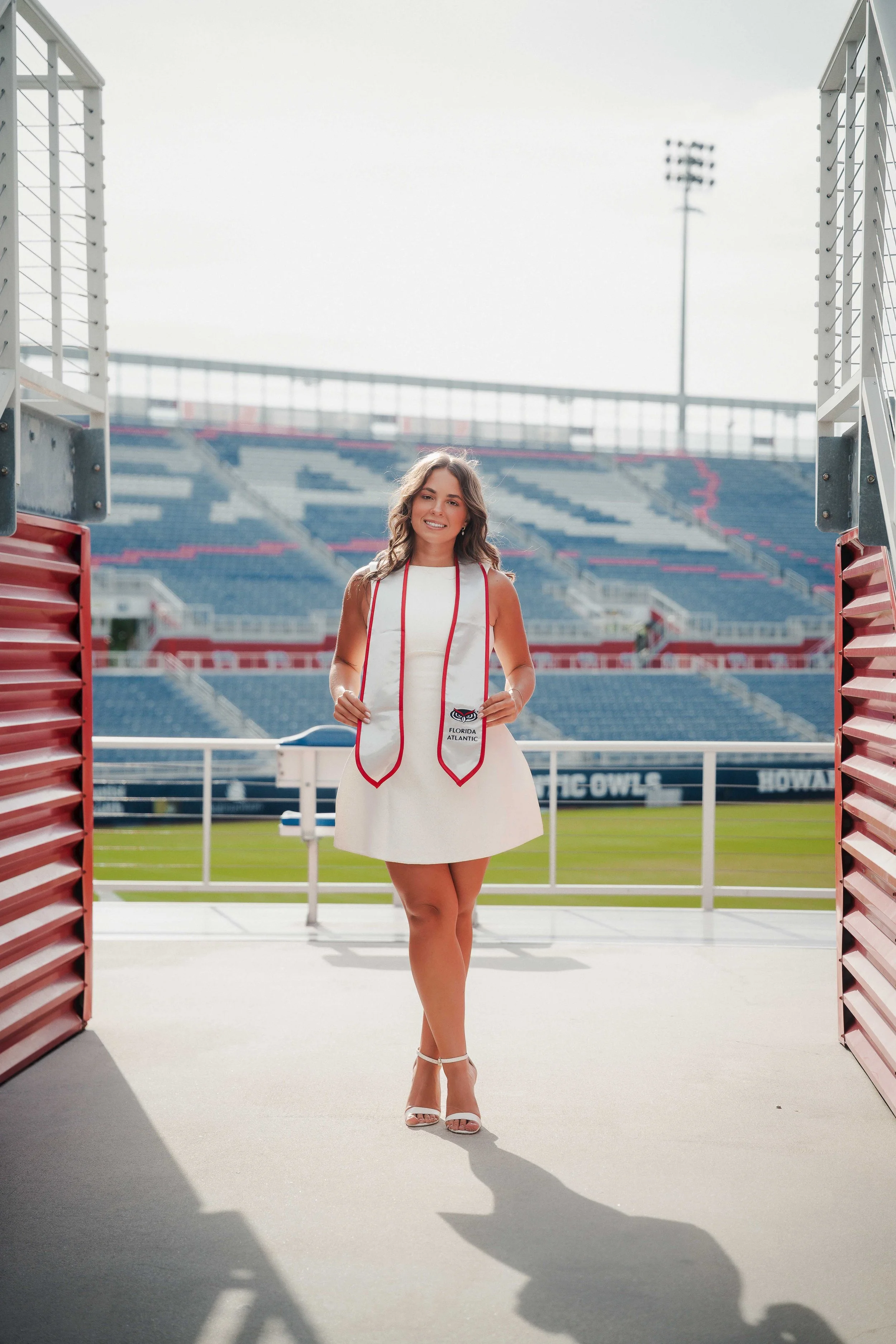 A woman in a white dress with red accents and high heels standing in stadium entrance, holding a stole with the Florida Atlantic University logo, with stadium seats and floodlights in the background.