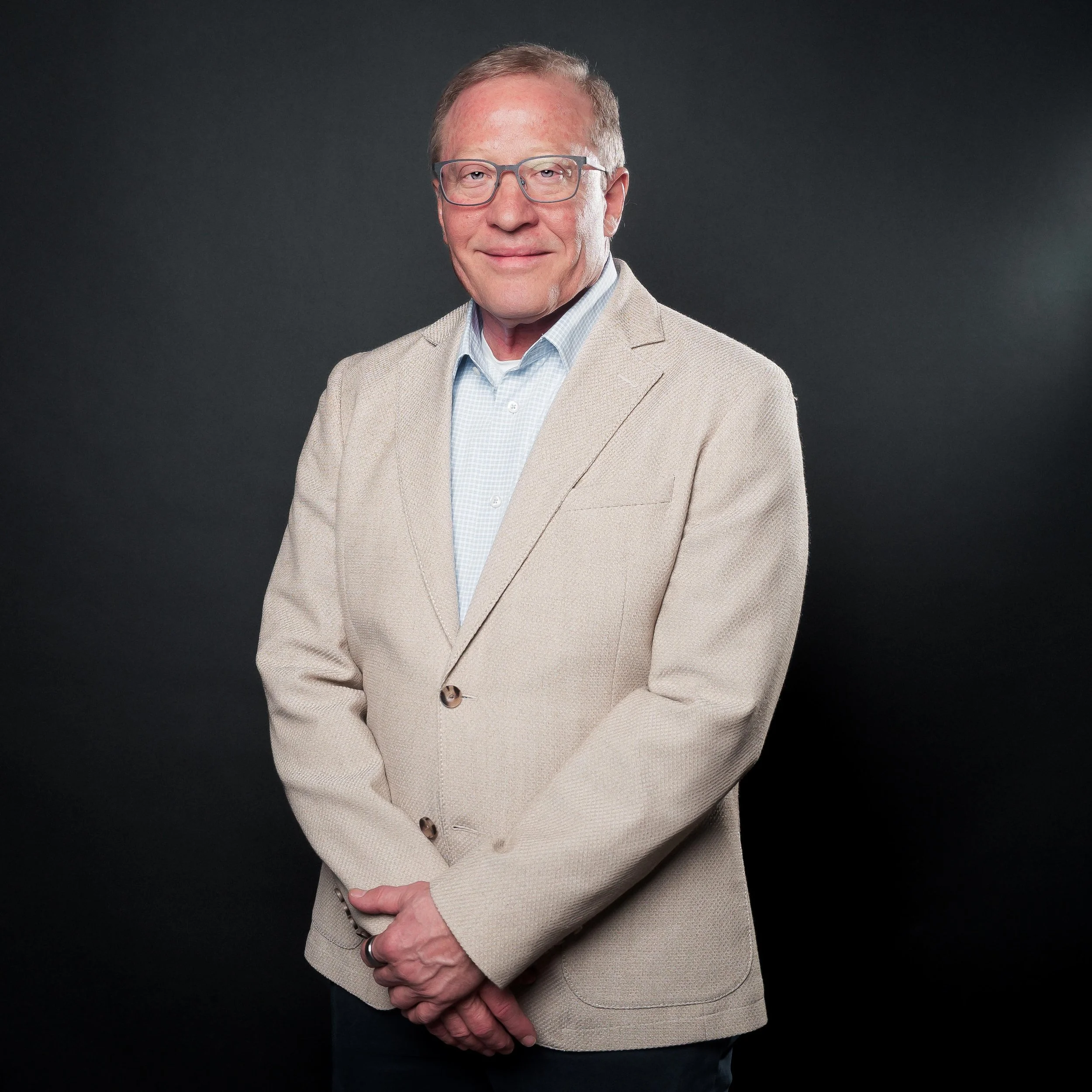 Close-up of an older man in a beige blazer and glasses, smiling against a dark background.
