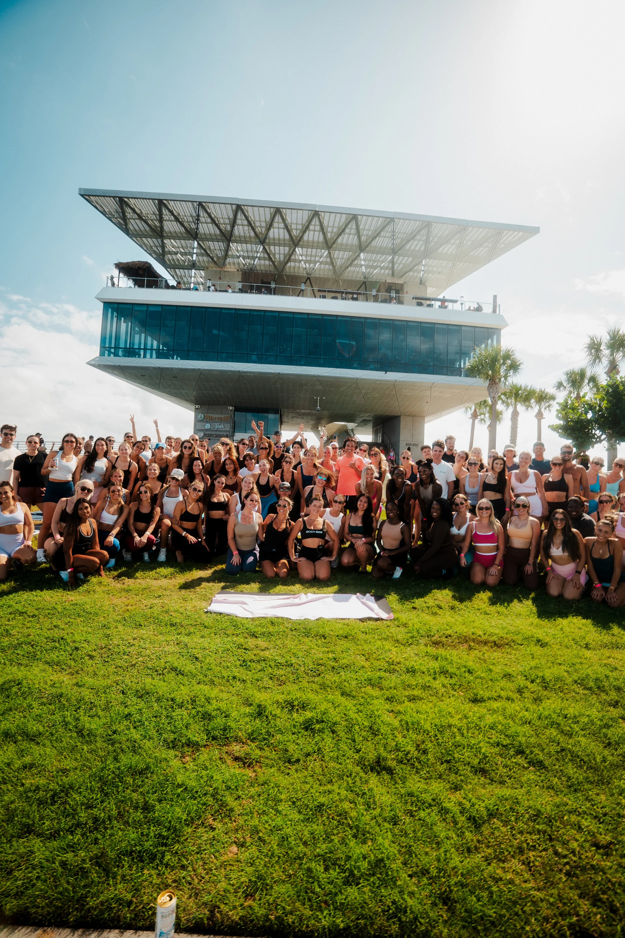 A large group of people gathered on a grassy area in front of a modern building with a distinctive architectural design and glass windows, with palm trees in the background.