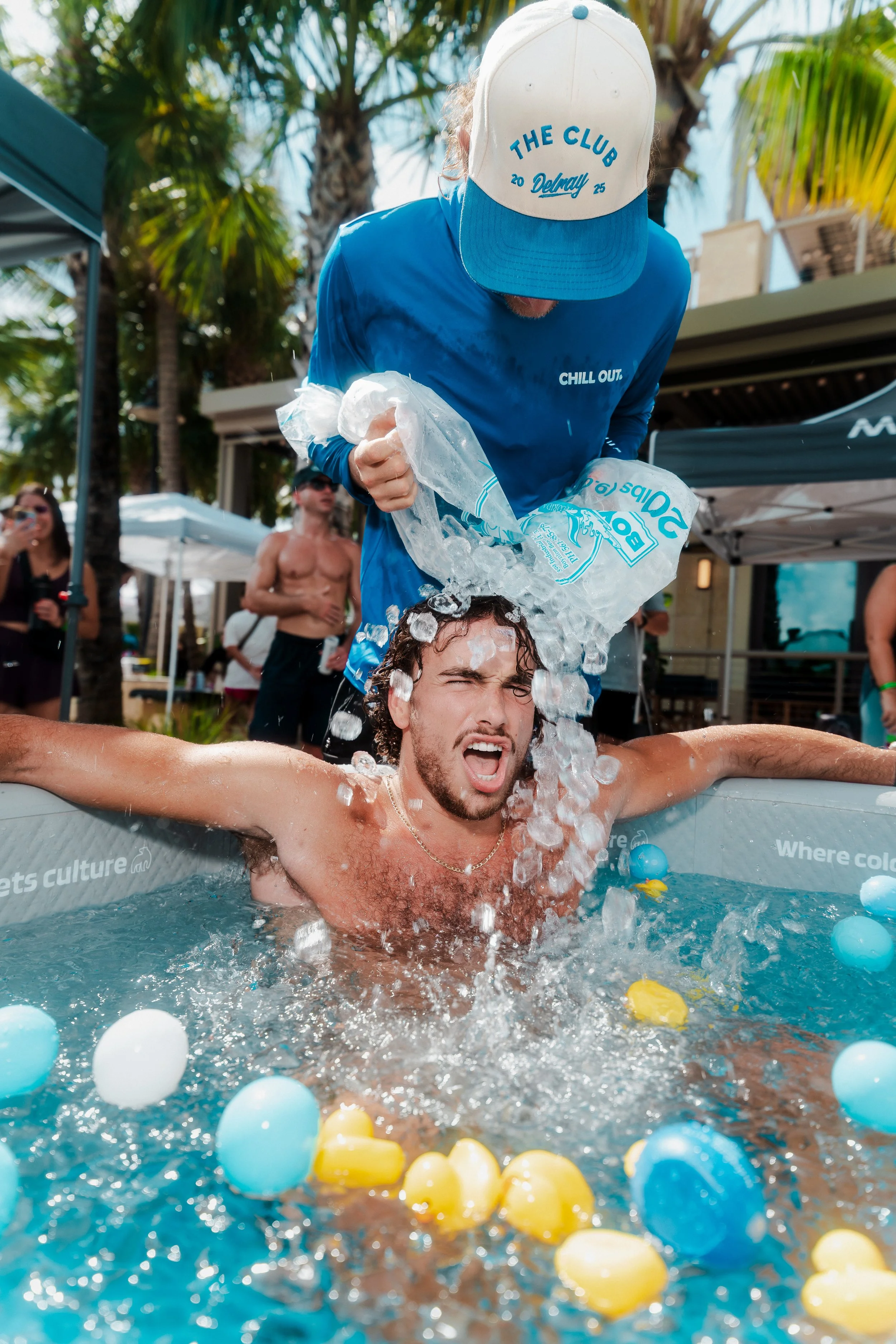 A man in a pool has his head over the water and is smiling with closed eyes, while a person standing behind him is pouring ice from a plastic bag onto his head. The scene is outdoors, with palm trees and tents visible in the background.