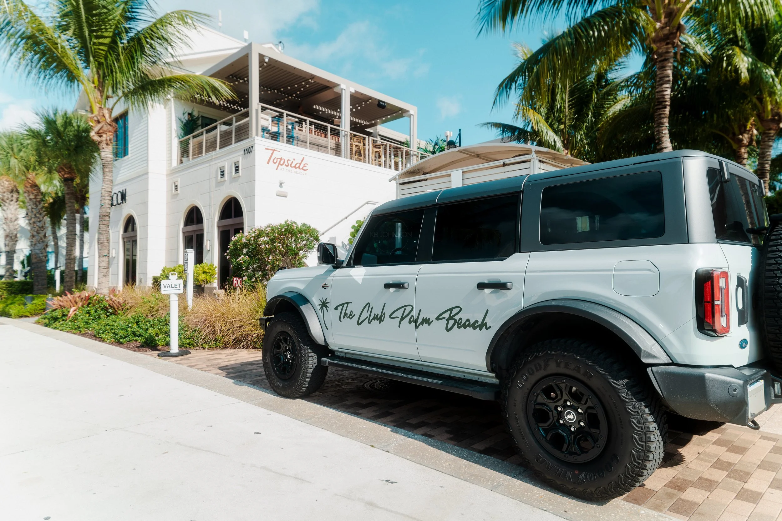 A white SUV parked on a street near a building with palm trees. The vehicle has the words 'The Club Palm Beach' on its side and is near a valet stand. The building has a sign that reads 'Topside at the Beach' and features an outdoor patio with string lights under a cloudy blue sky.