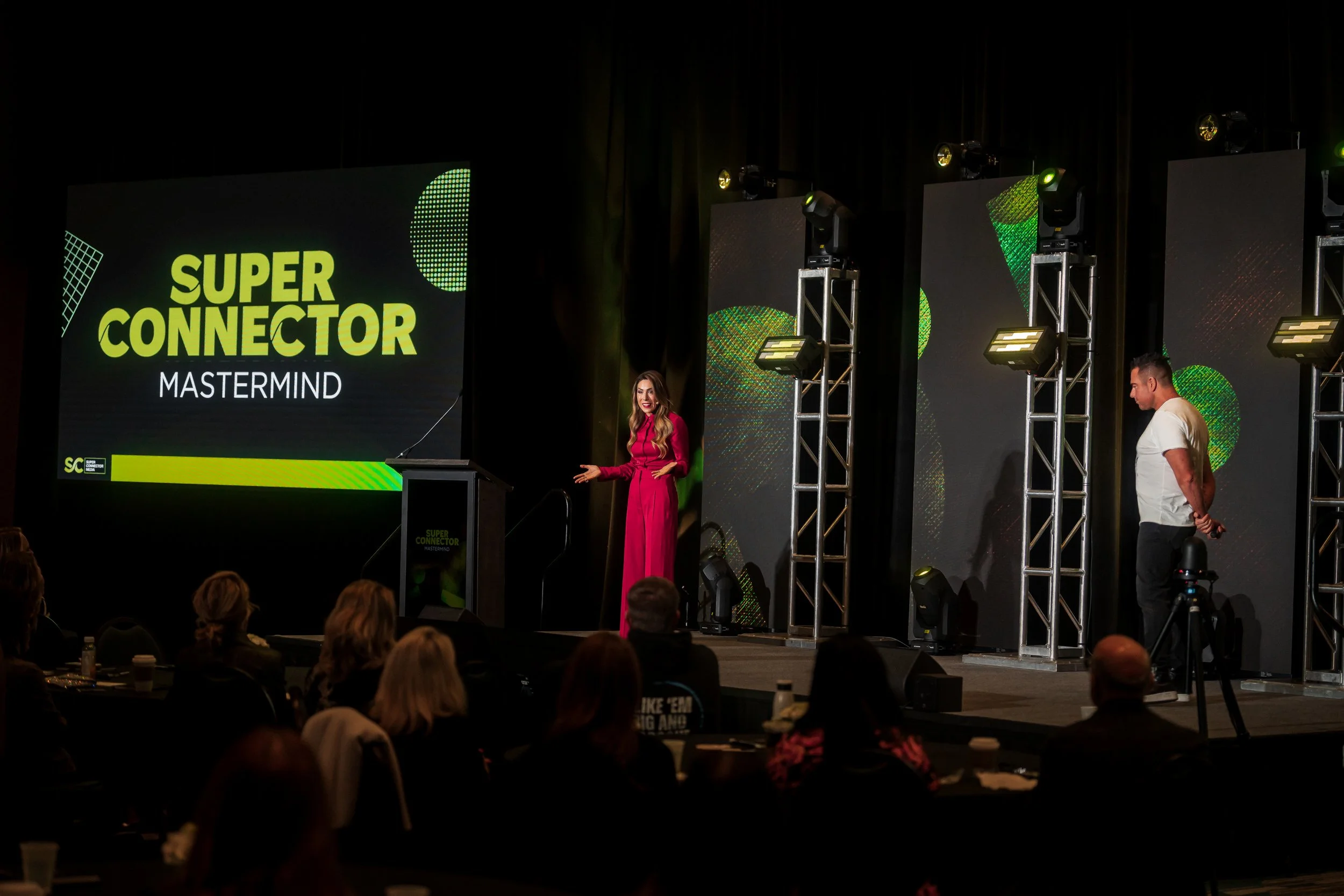 A woman in a pink dress speaking at a stage with a large screen that reads "Super Connector Mastermind". Several people are seated in the audience, and stage lighting is visible.