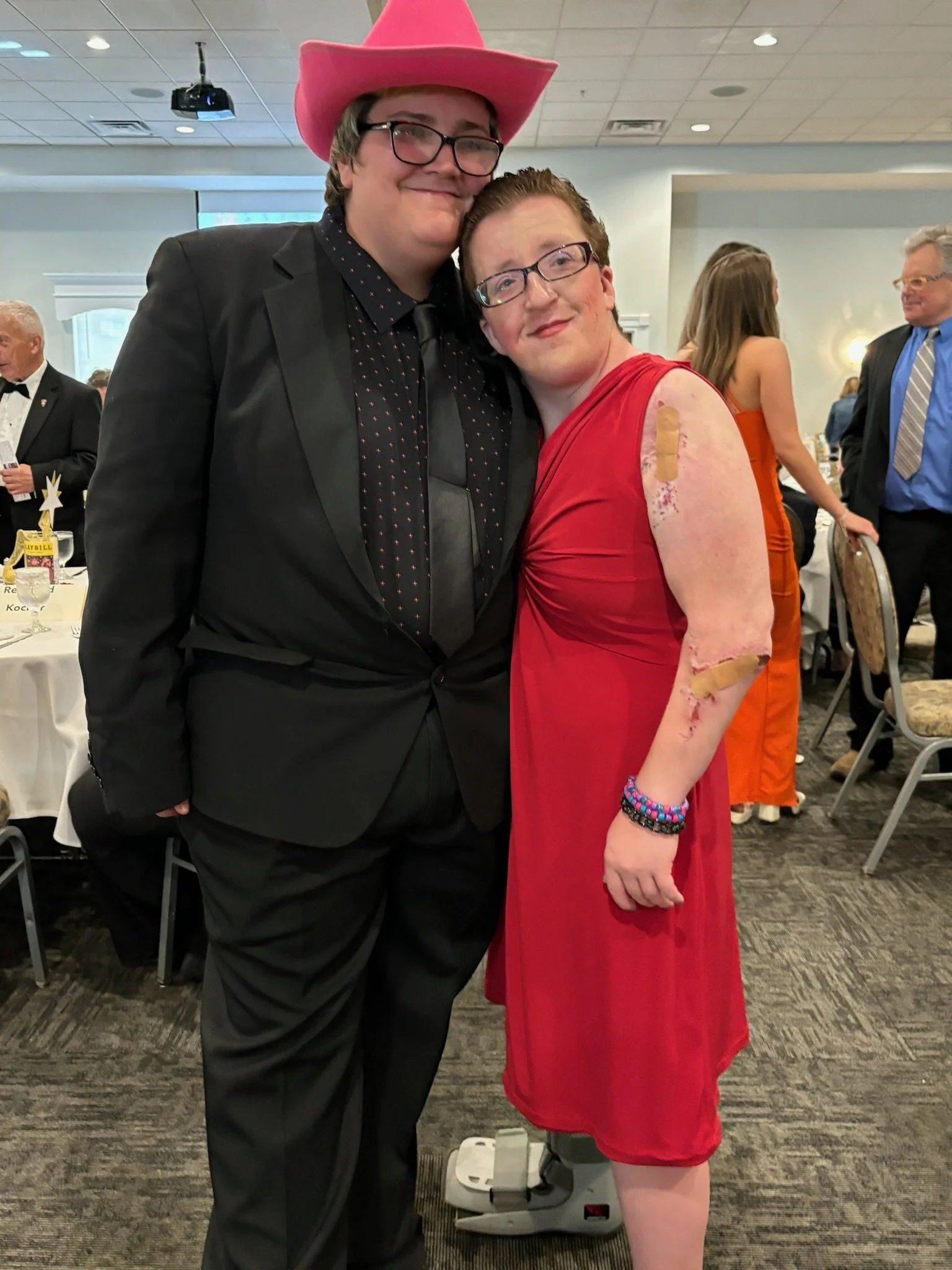 A young man in a black tuxedo and pink cowboy hat standing next to a young woman in a red dress with visible scars and bandages on her arms, at a formal event.