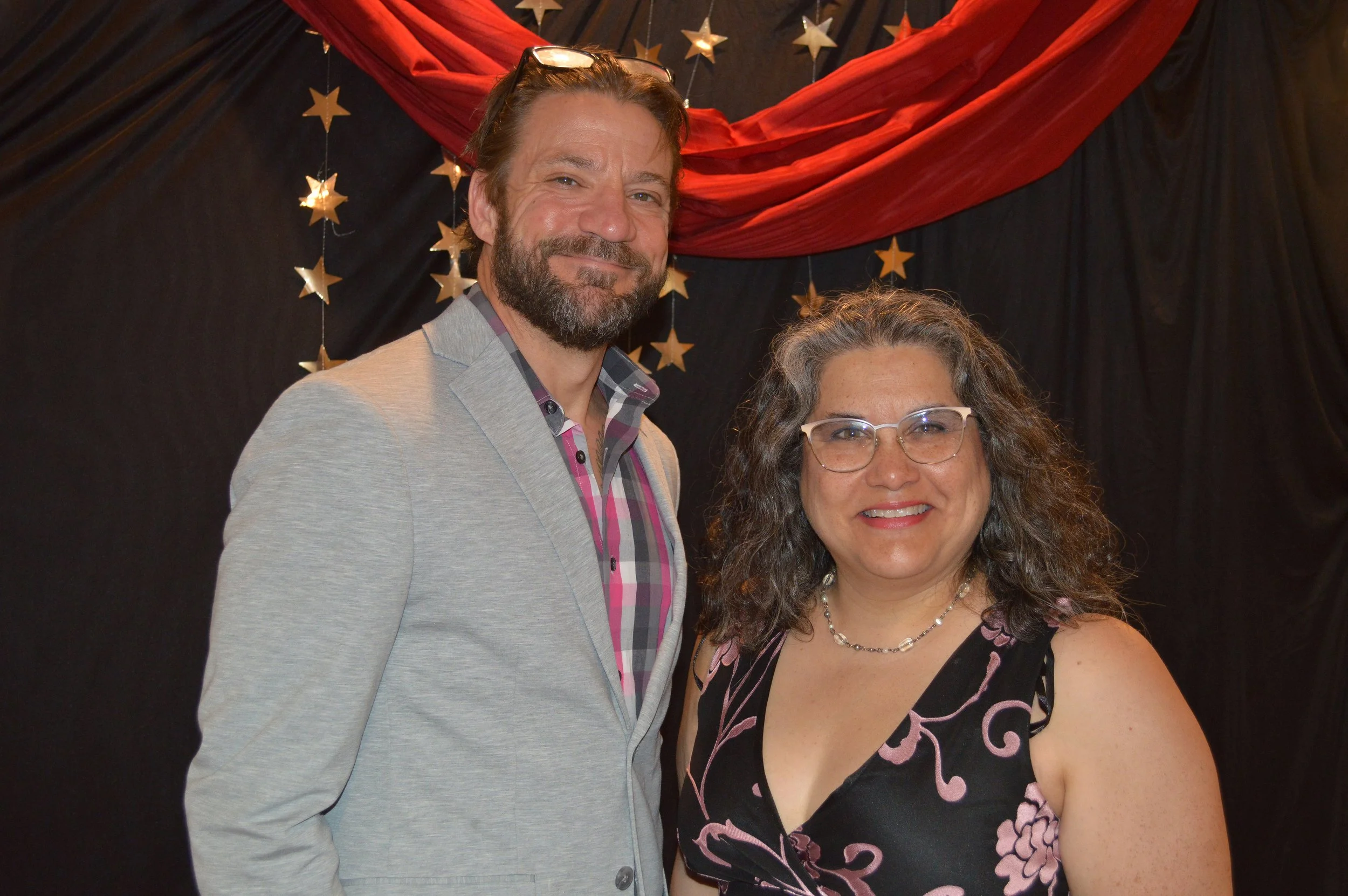 A man and a woman pose smiling in front of a backdrop with black fabric, red drapery, and gold star decorations.