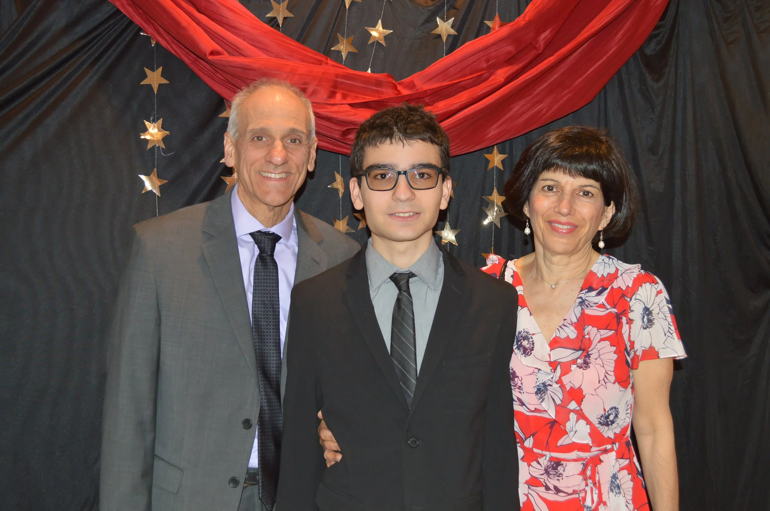 Three people, two men and one woman, are standing in front of a black backdrop with gold star decorations and red fabric. The man on the left is wearing a gray suit, white shirt, and black tie. The young man in the middle has dark hair, glasses, and 
