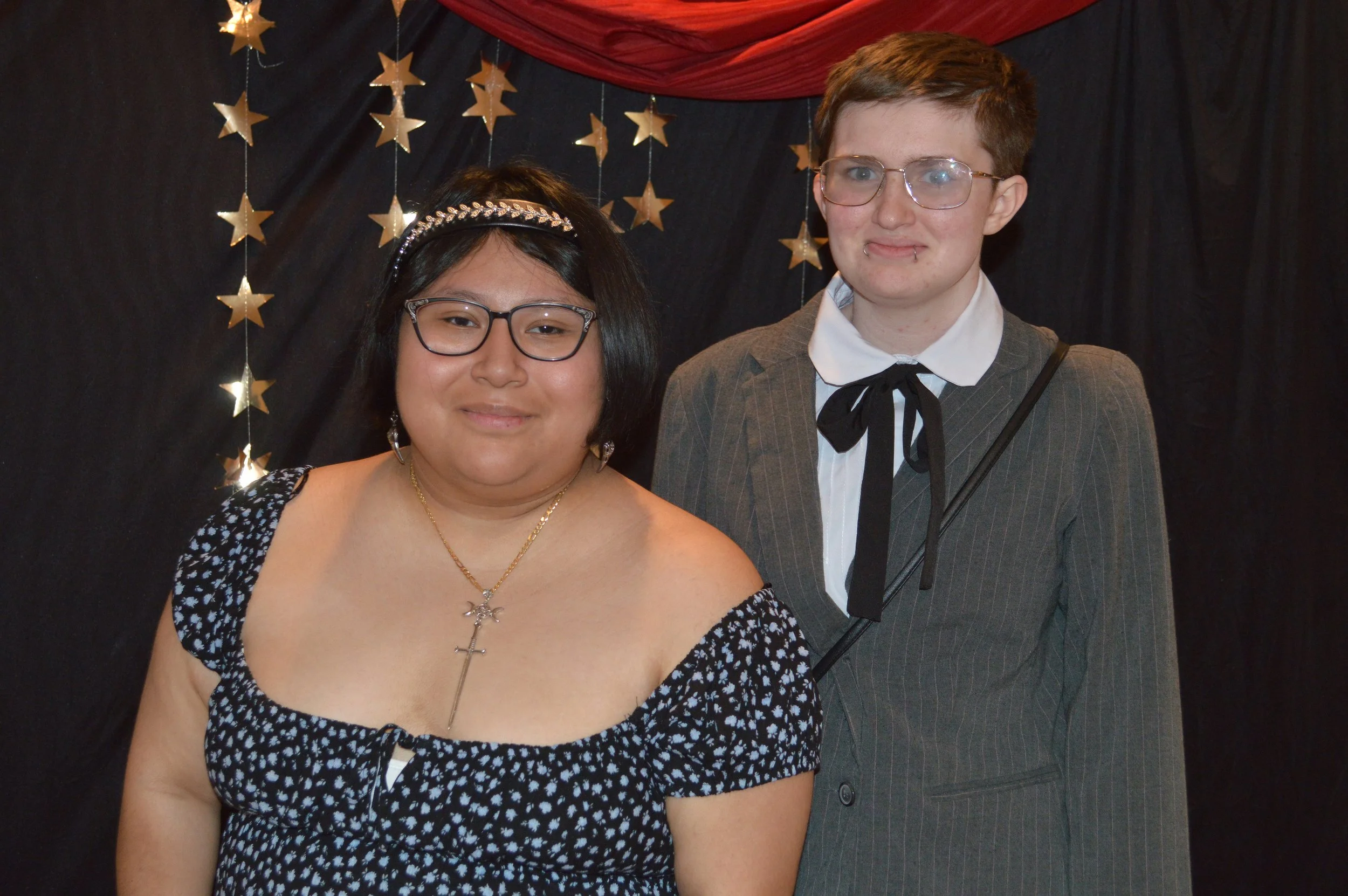 Two women standing in front of a black backdrop decorated with gold star-shaped hangings. One woman has short dark hair, glasses, and is wearing a black and white polka dot dress with a square neckline, jewelry, and a headband. The other woman has sh