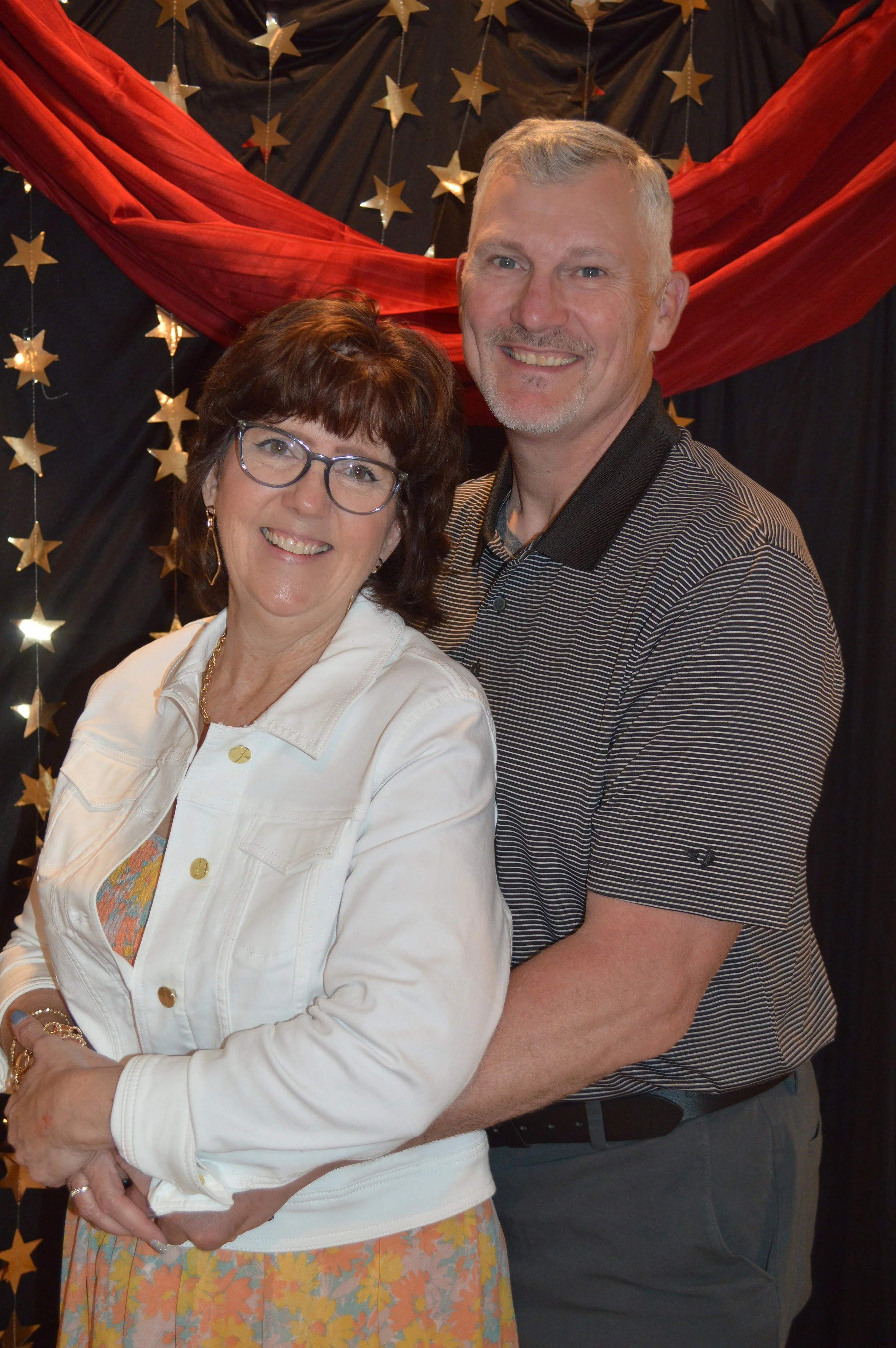 A smiling couple back-to-back, with a backdrop of gold stars and red drapery, at a festive event or celebration.