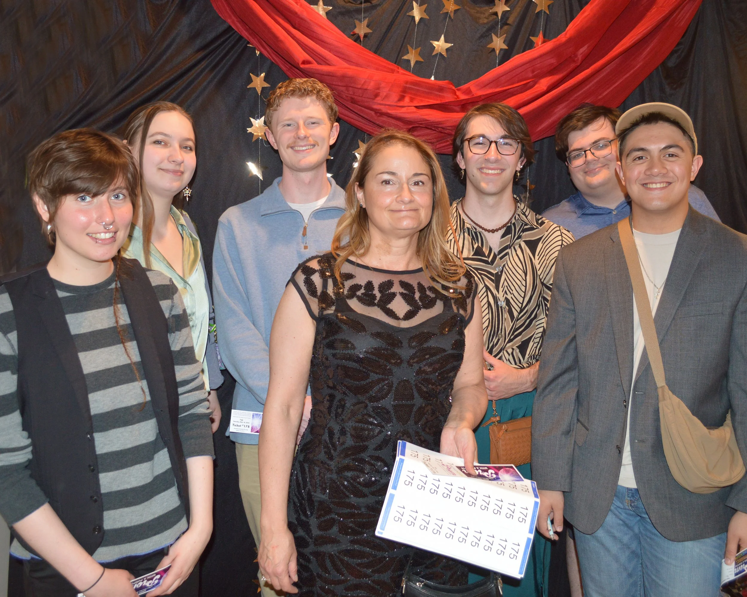 Group of seven people standing together, including a woman holding a stack of tickets, in front of a decorative backdrop with stars and red drapery.
