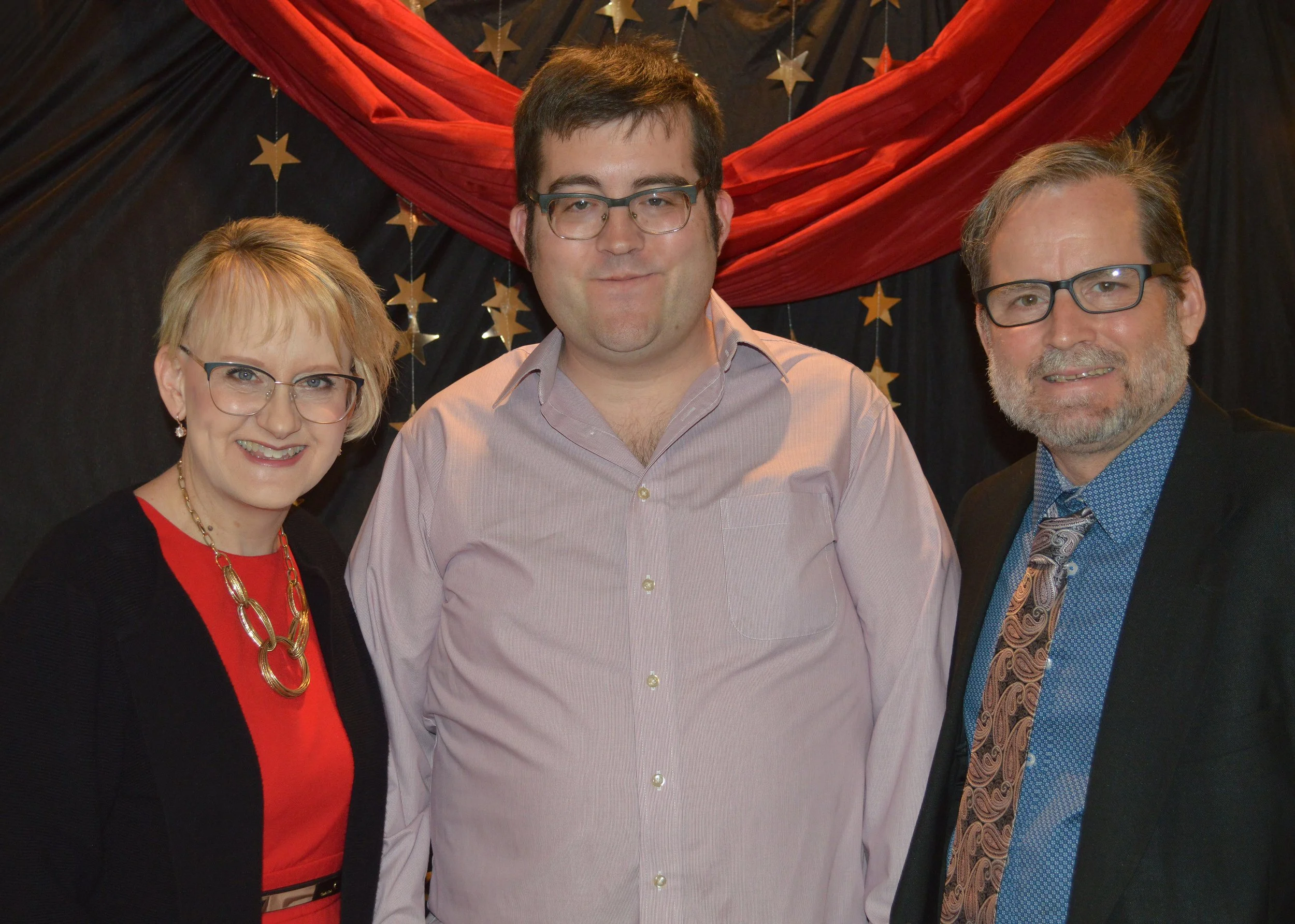 Three people, two men and one woman, standing close together indoors, with a background of black fabric decorated with gold stars and red fabric drapery.