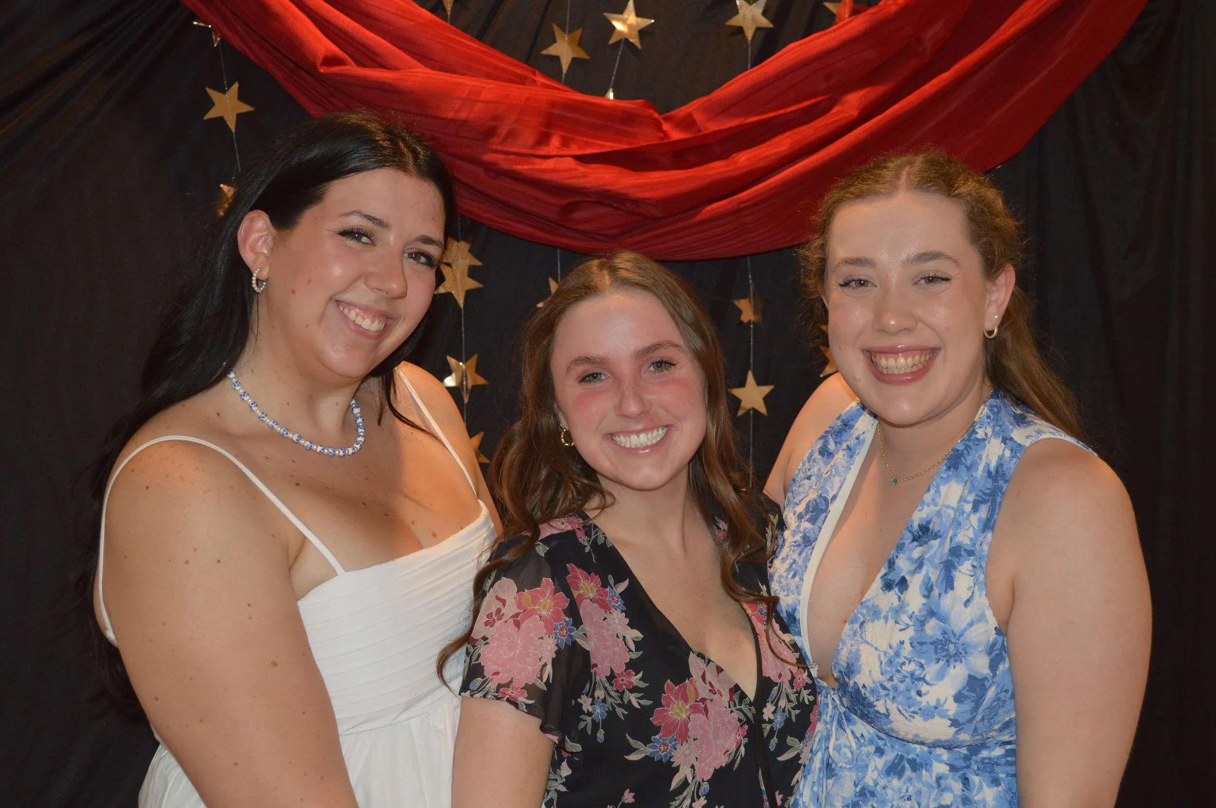 Three women smiling, standing close together in front of a black backdrop with gold star decorations and red fabric drapery.