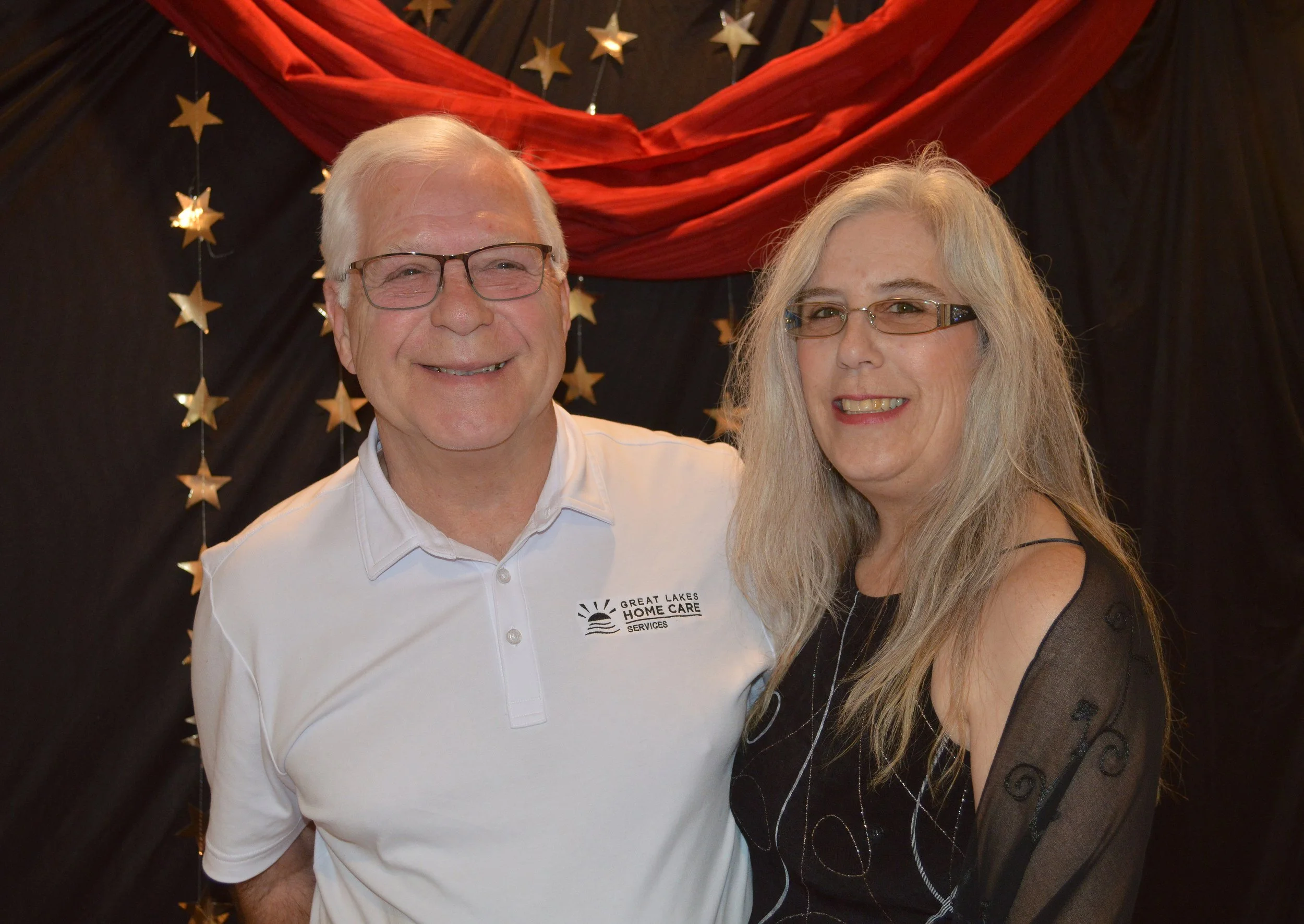 A smiling elderly man in glasses wearing a white shirt with a logo that reads 'Great Lakes Home Care Services,' standing next to a smiling woman with long gray hair, glasses, and wearing a black top with sheer patterned sleeves, in front of a backdro