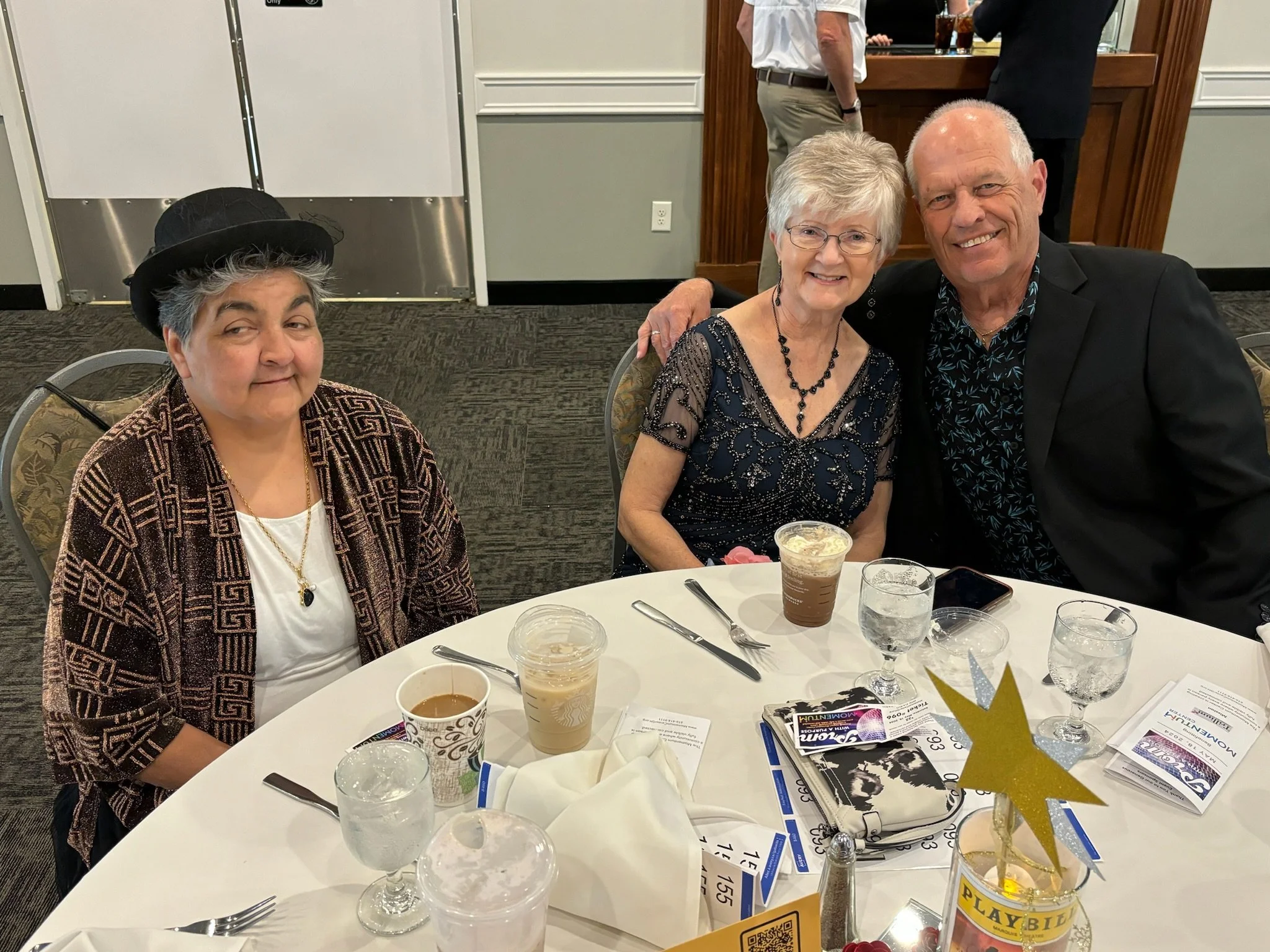 Three elderly people sitting at a round table during a celebration or event, with two women and one man smiling, and a woman wearing a black hat and a patterned jacket.