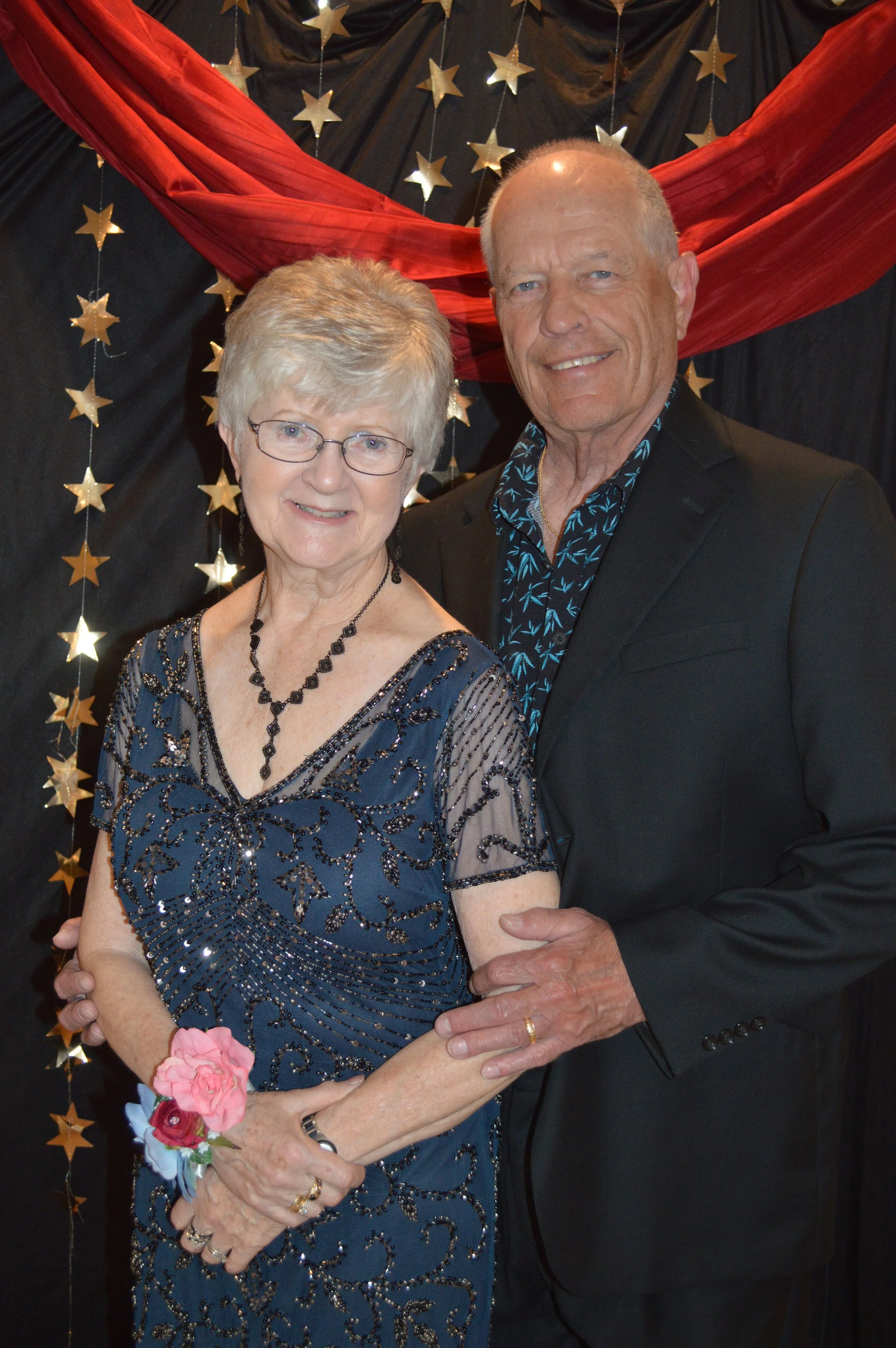 Older woman and man dressed in formal attire, standing close together and smiling at the camera, with a backdrop decorated with black fabric, gold star decorations, and red drapery.