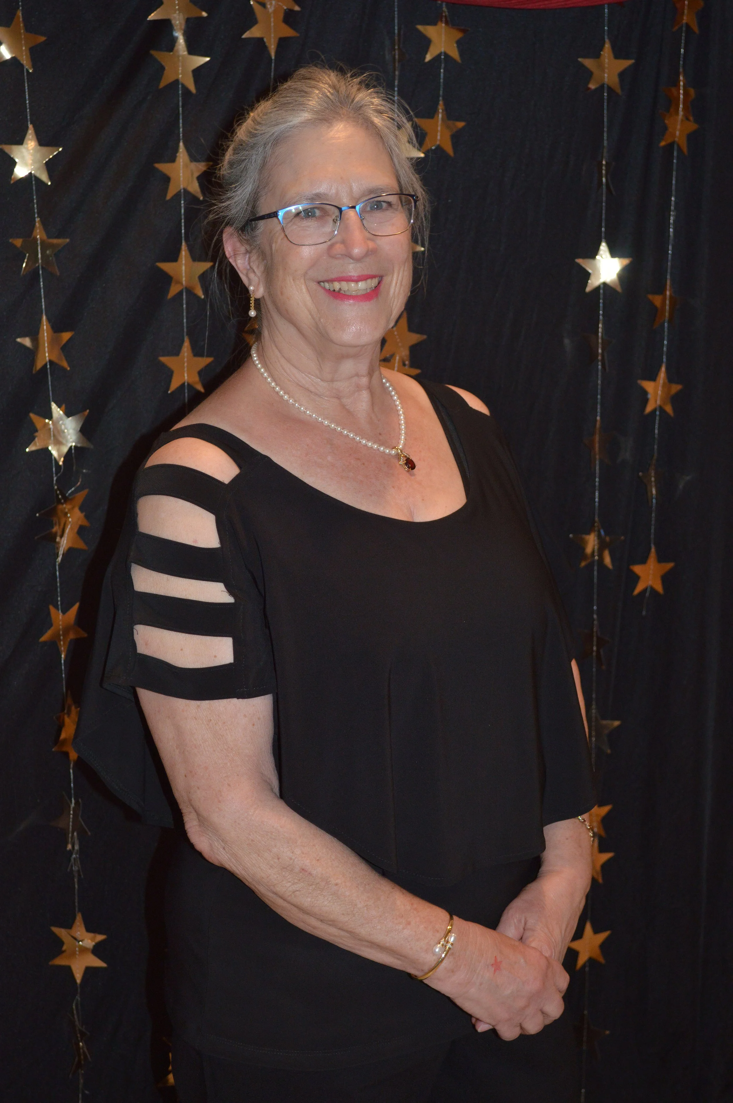 An elderly woman with glasses and gray hair, wearing a black top with cut-out shoulders, a pearl necklace, earrings, and bracelets, stands in front of a black backdrop decorated with gold star garlands, smiling at the camera.