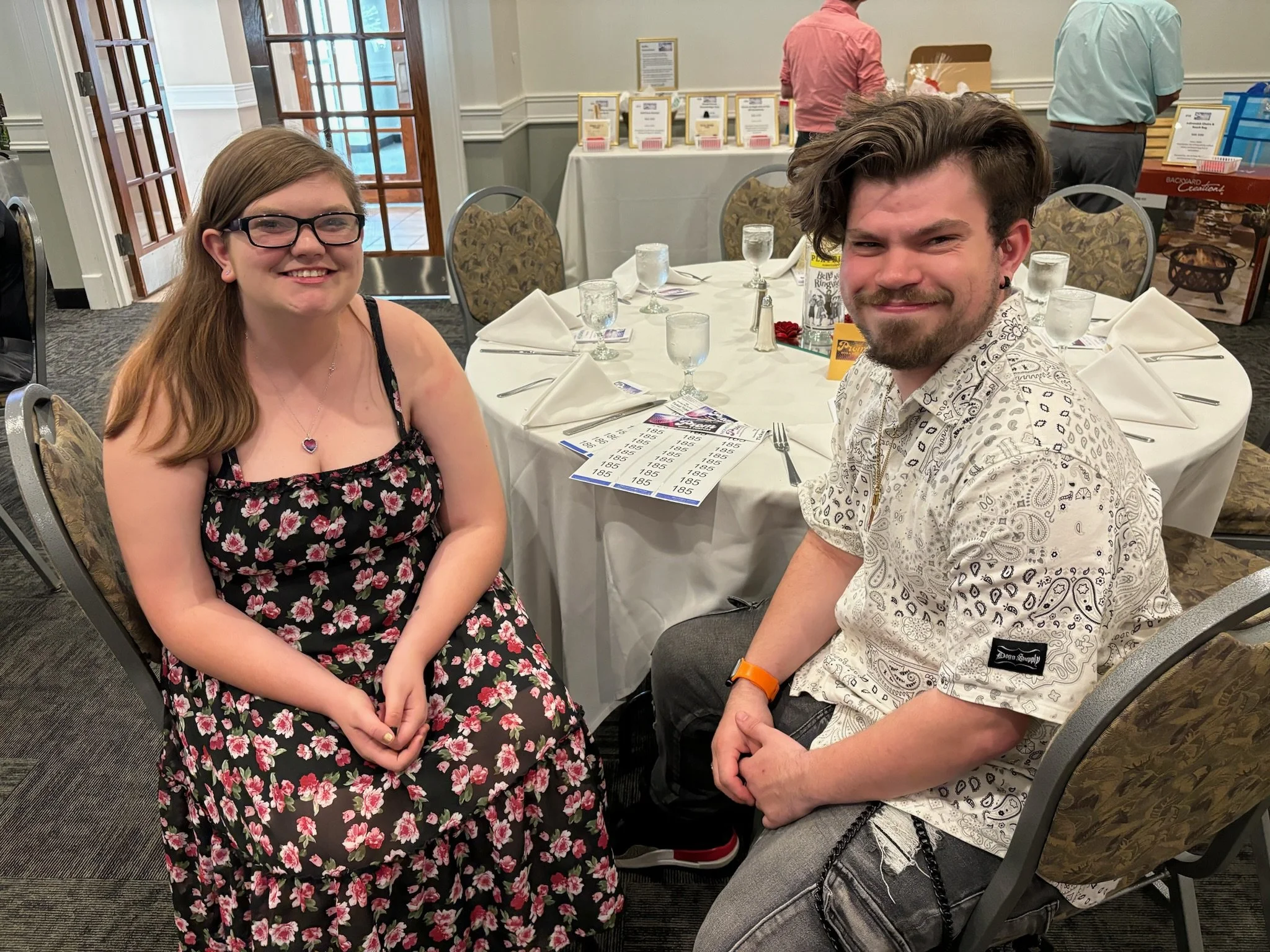 Two smiling people sit at a round table in a banquet hall, with one woman wearing glasses and a floral dress, and a man with a beard and casual patterned shirt.