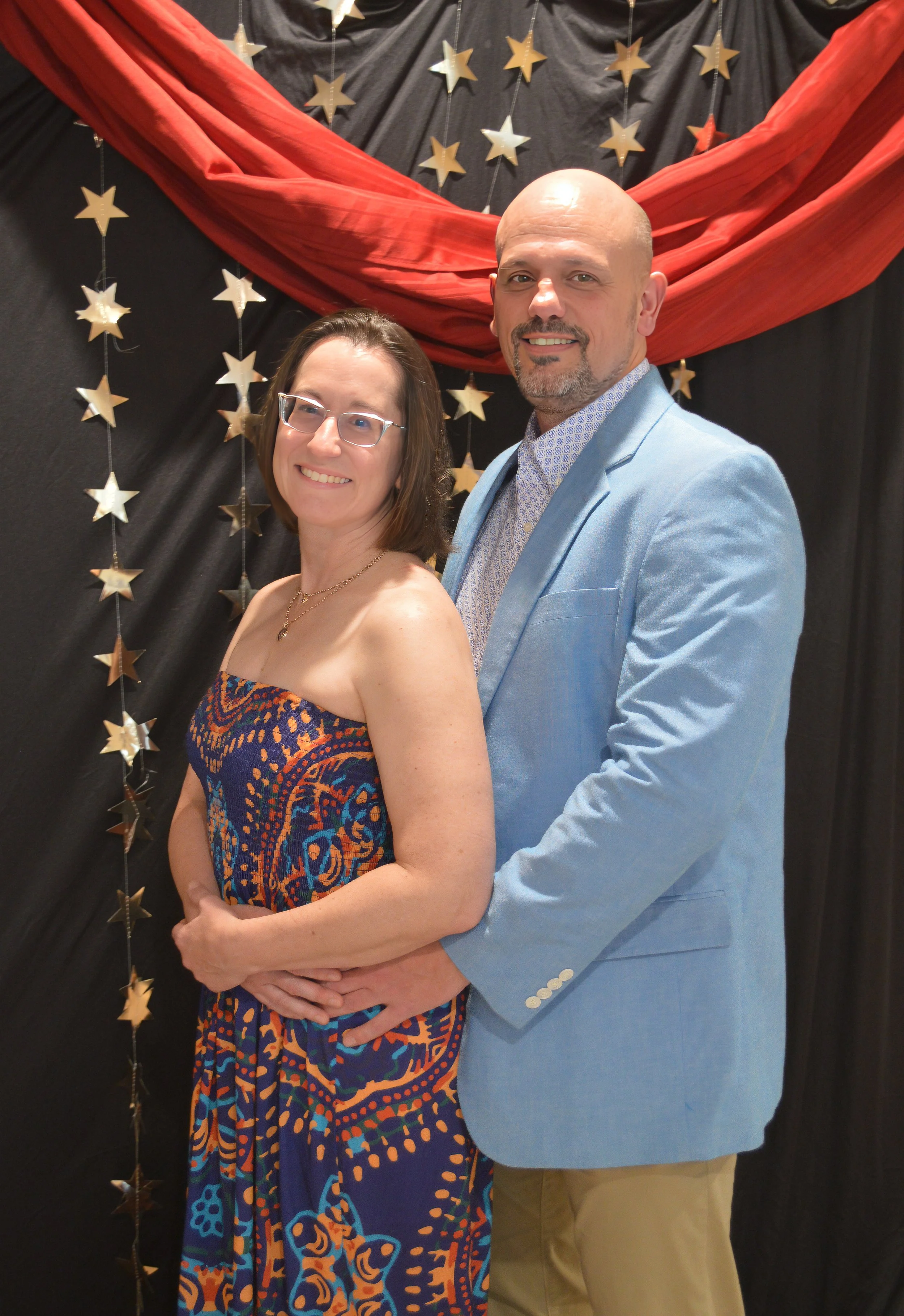 A smiling couple stands together in front of a black backdrop with gold star decorations and a red fabric draped overhead. The woman wears glasses and a strapless, colorful patterned dress, while the man wears a light blue blazer, a patterned shirt, 
