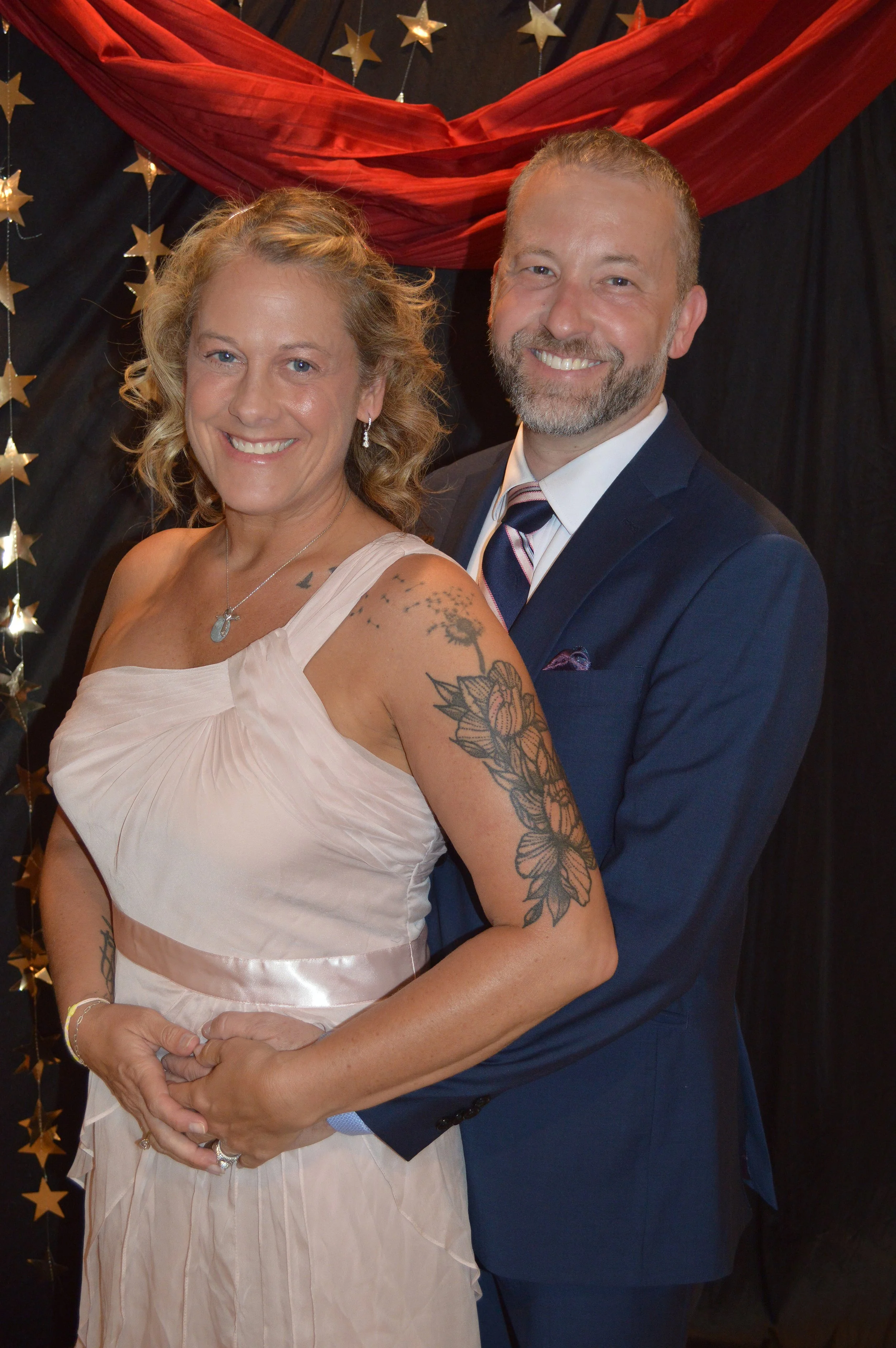 A smiling woman in a light pink dress with tattoos on her arm, and a man in a navy blue suit with a striped tie, pose together at a decorated event with black curtains, red fabric, and gold star decorations in the background.